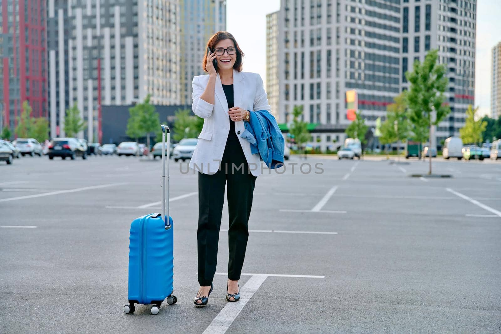 Middle aged businesswoman with suitcase talking on mobile phone in outdoor parking, urban style, city architecture background. Business travel trip, business people of mature age, success, modern city