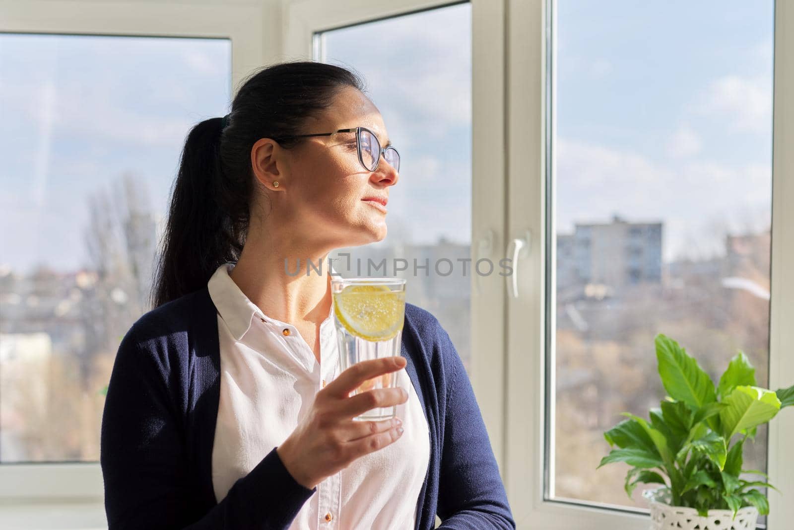 Business woman with glass of water with lemon. Middle-aged female in glasses, white shirt cardigan near window, resting enjoying healthy vitamin water