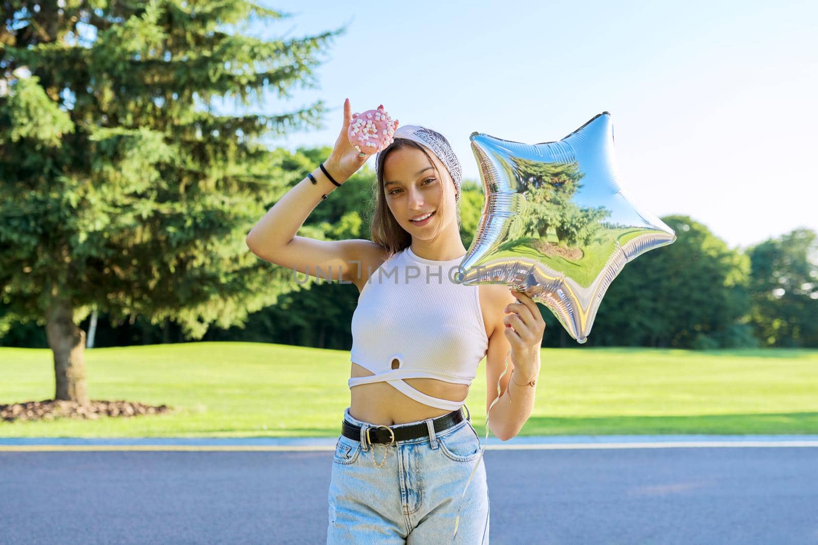 Fashionable beautiful girl teenager with sweet donut and balloon star in her hands, sunny summer day, park, road background. Youth, teens, summer, fashion, street style, lifestyle concept