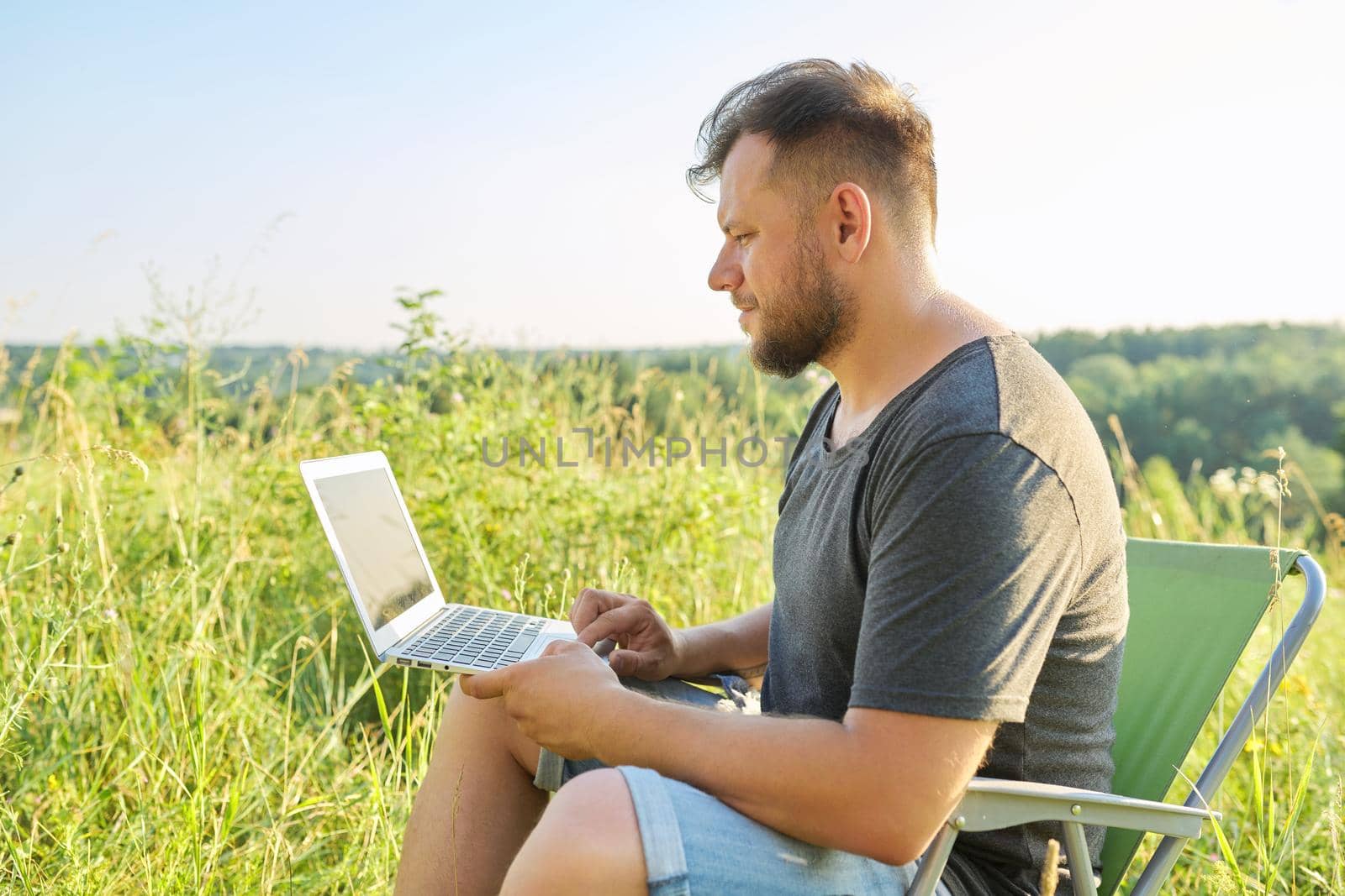Middle-aged man with laptop in nature. Bearded male sitting on an outdoor chair in meadow in nature, enjoying summer day and using modern technologies for rest and work