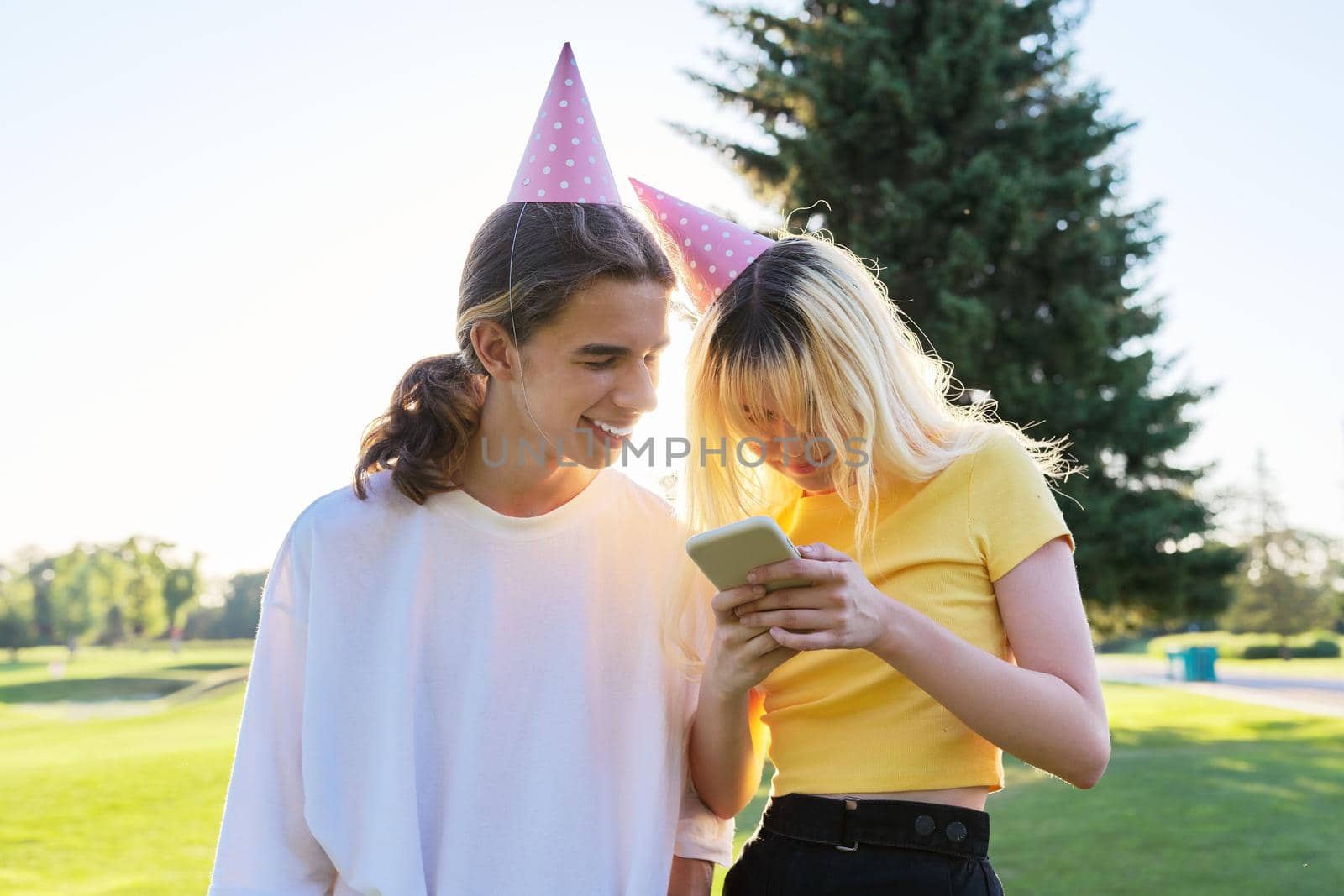Birthday party, happy couple of teenagers boy and girl 16, 17 years old in festival hats with smartphone outdoor