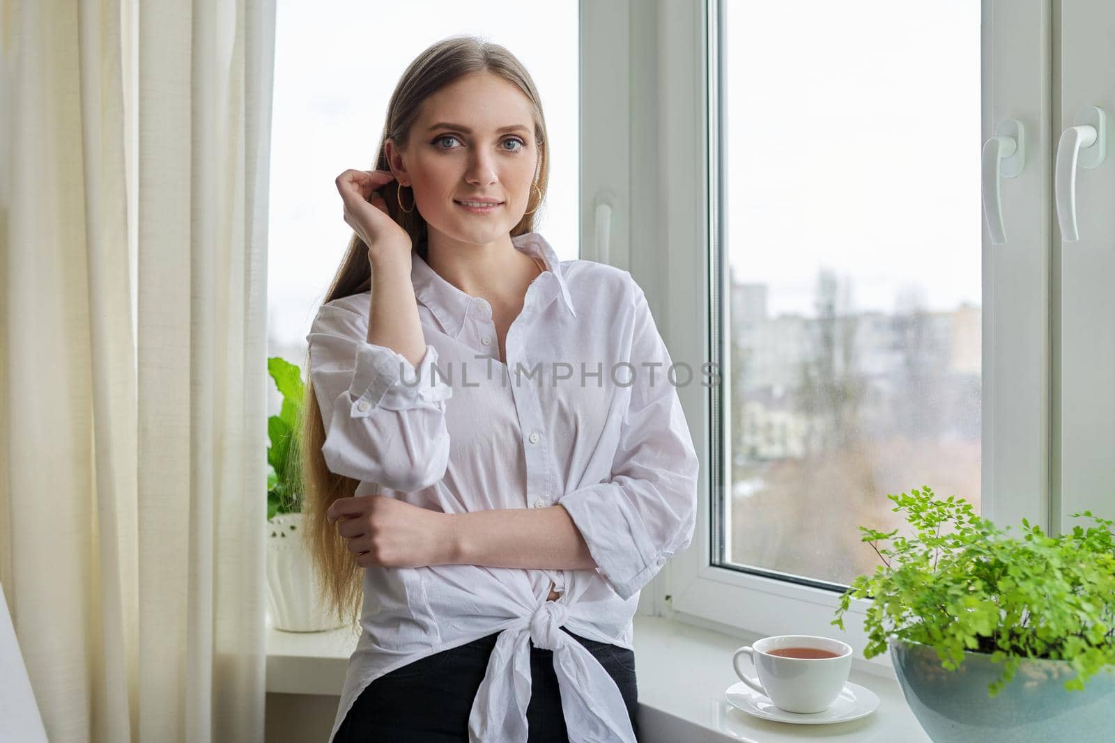 Portrait of young beautiful blonde woman in white shirt near winter autumn window. Smiling 20s blonde female relaxing looking at camera
