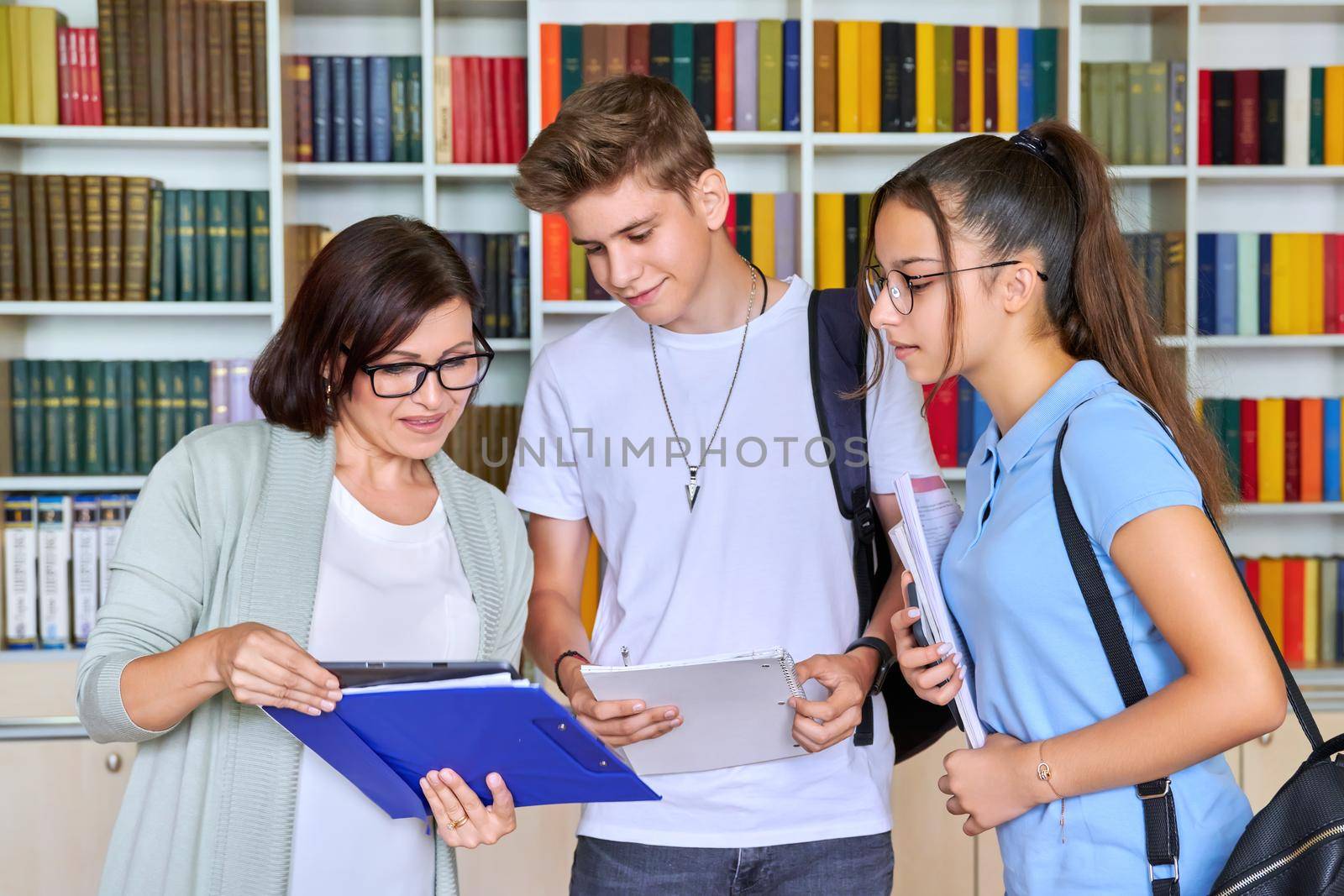 Students teenagers talking with woman teacher mentor, library shelving with books background. High school, education, knowledge, adolescence concept