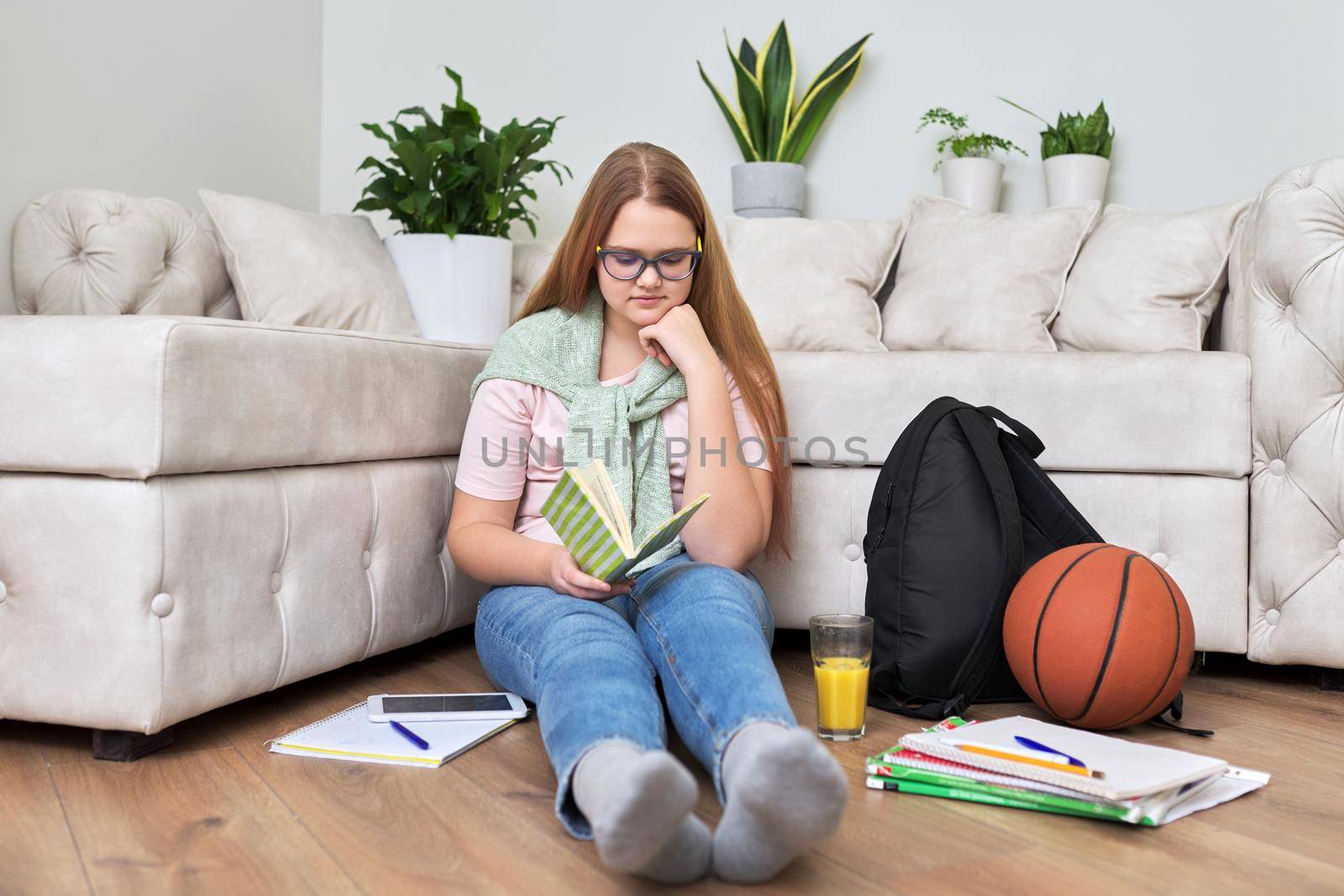 Lifestyle, education children 12, 13 years old concept. Teenage girl sitting at home on the living room floor reading book with backpack, books, school notebooks, basketball