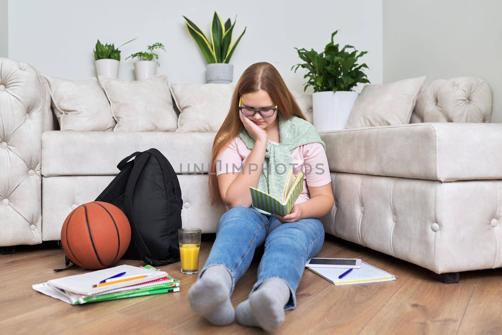 Lifestyle, education children 12, 13 years old concept. Teenage girl sitting at home on the living room floor reading book with backpack, books, school notebooks, basketball