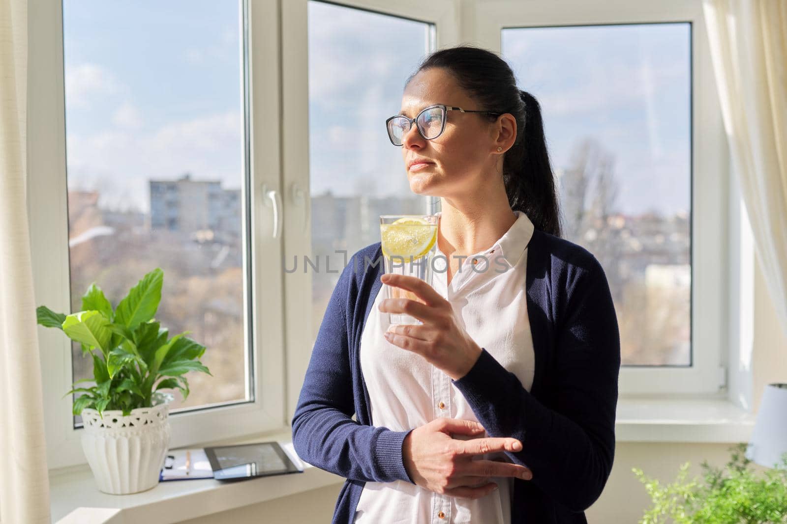 Business woman with glass of water with lemon. Middle-aged female in glasses, white shirt cardigan near window, resting enjoying healthy vitamin water