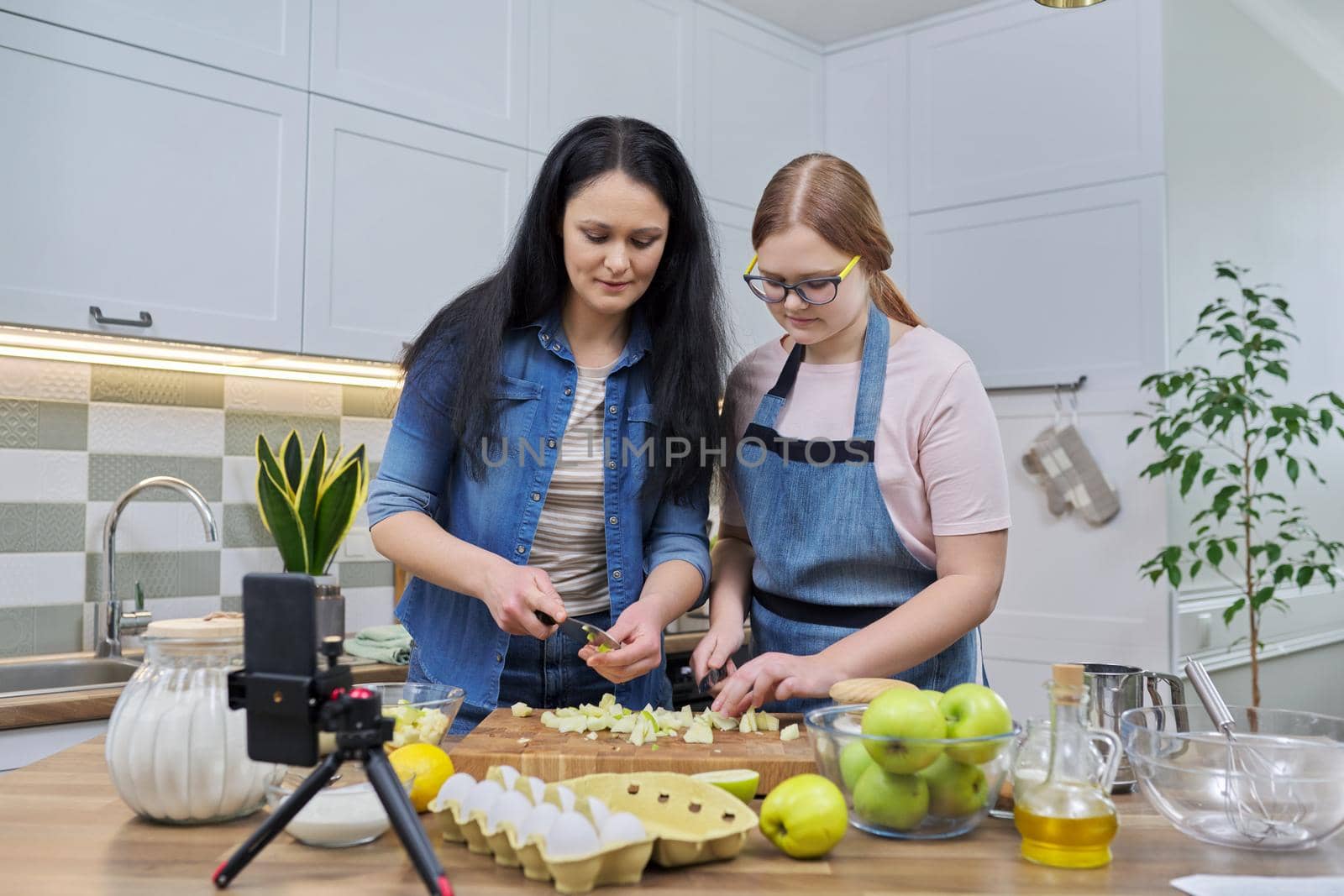 Mom and teen daughter cooking apple pie together, looking at smartphone screen, video recipe, writing recipe on family food blog, talking online using video communication