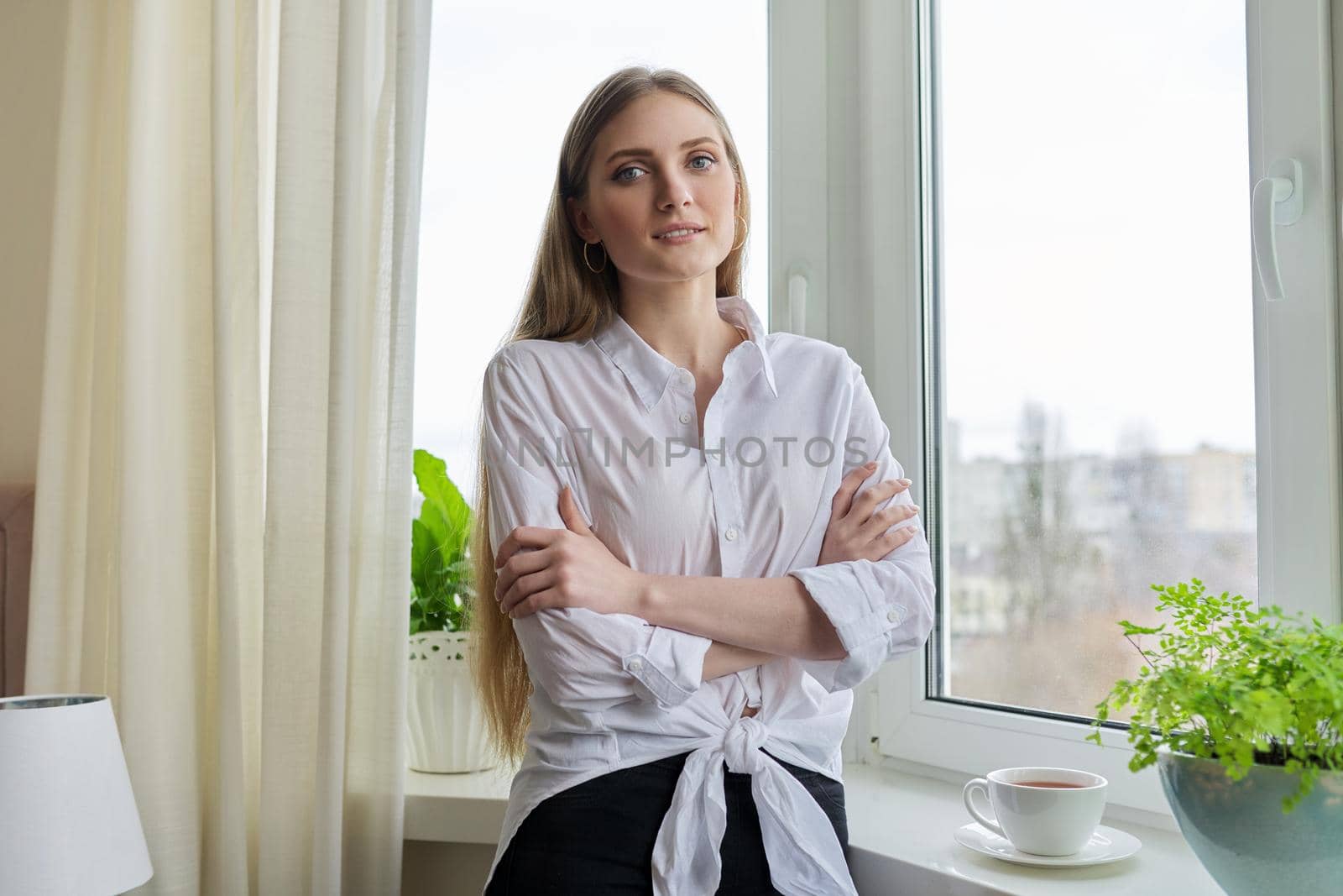 Portrait of young beautiful blonde woman in white shirt near winter autumn window. Smiling 20s blonde female relaxing looking at camera