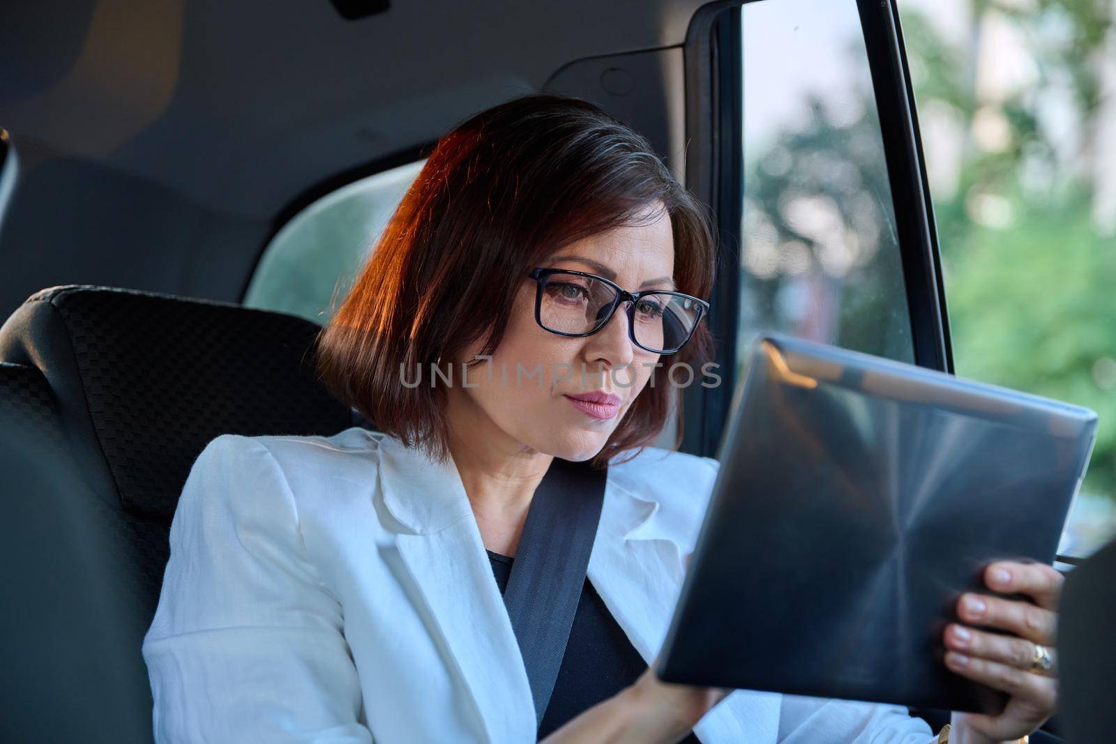 Serious middle-aged business woman in car in passenger backseat. by VH-studio