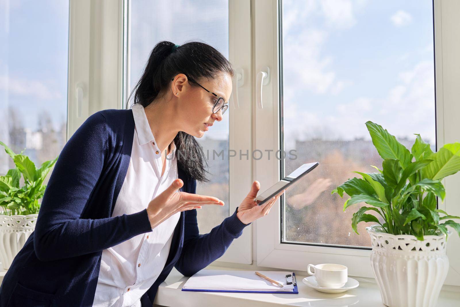 Serious business woman working remotely in her home office, talking on video call, using digital tablet. Freelance, technology, telecommunications, business remotely, video communication near window