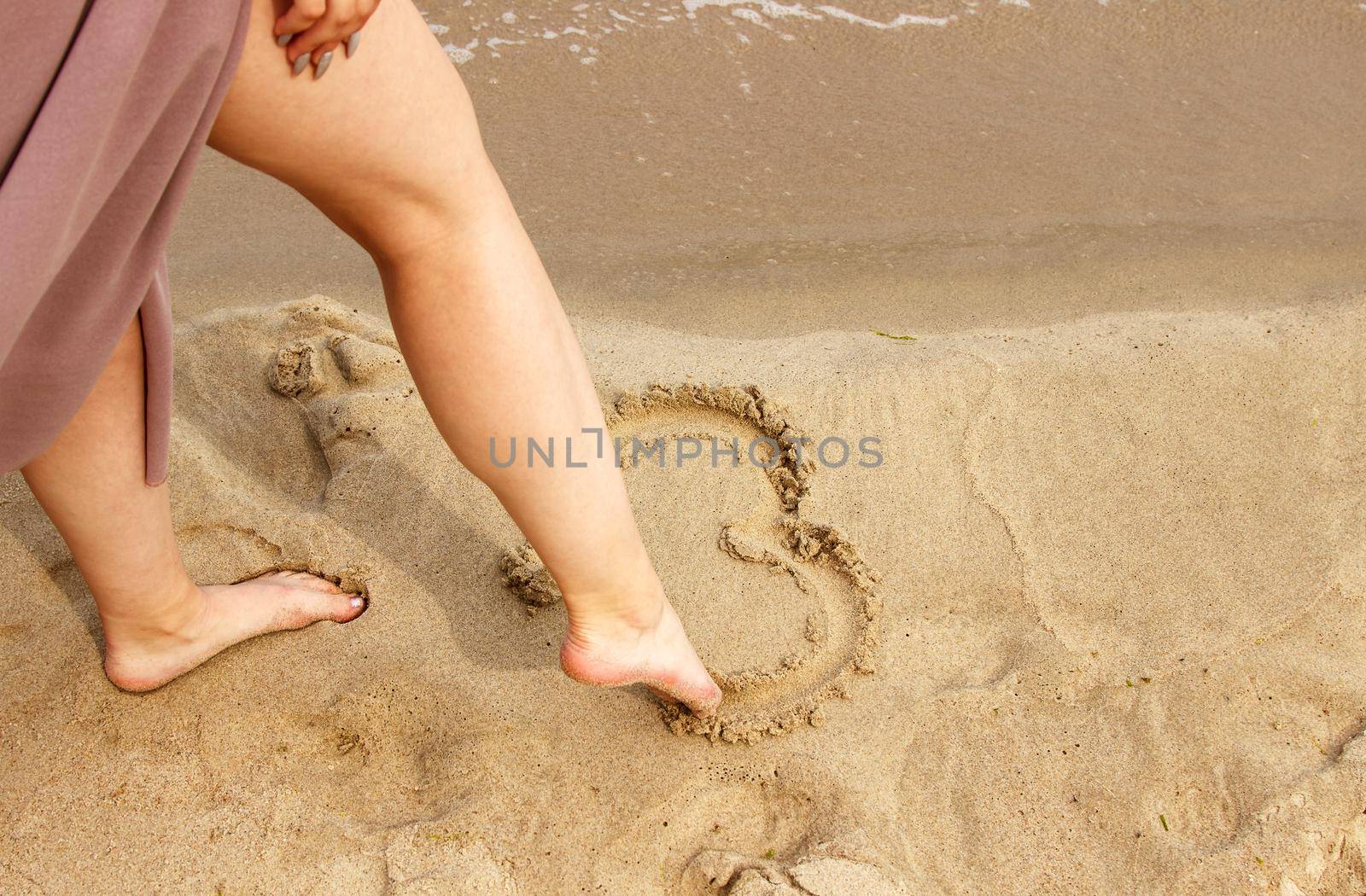 woman in beige dress draws with her foot heart on the sand by sea by raddnatt