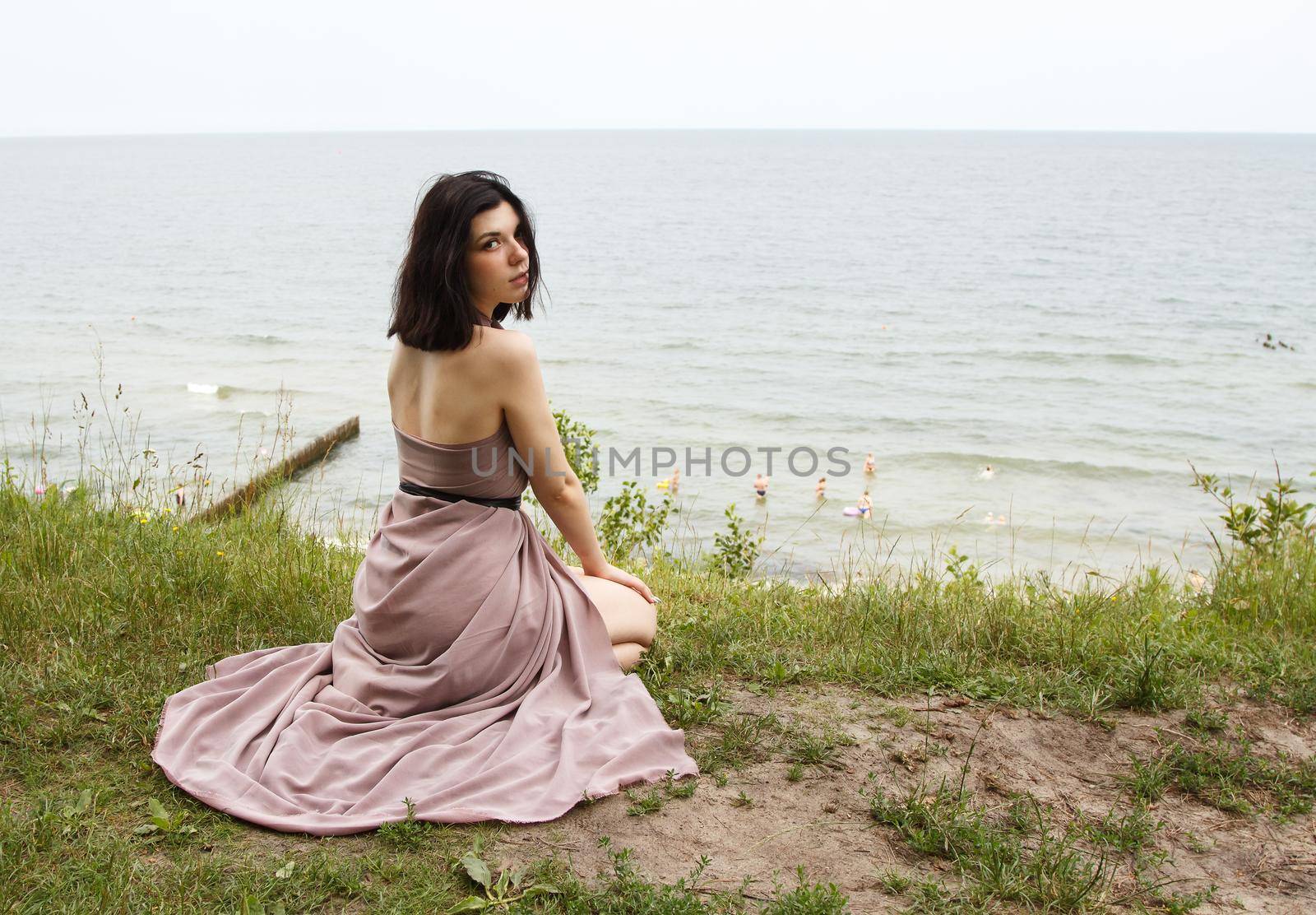 young brunette woman in beige dress sitting by the sea on summer day