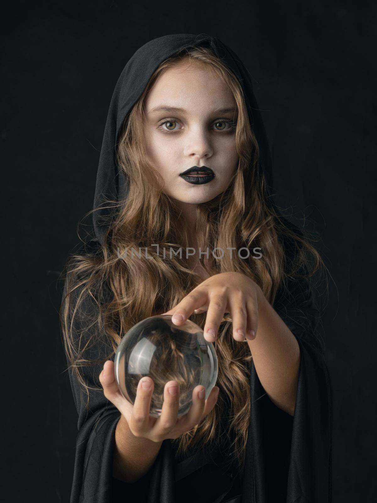 Little girl in Halloween witch costume and dark make-up holding crystal ball isolated on white background