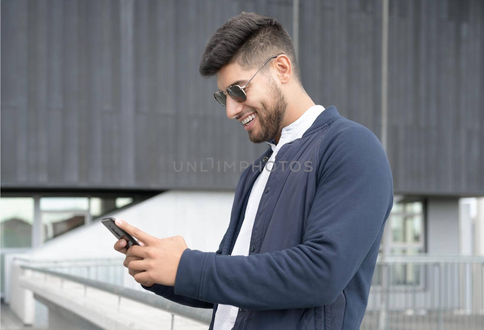 Handsome man using mobile phone app texting outside of office in urban city with skyscrapers buildings in the background. Young man holding smartphone for business work. by HERRAEZ