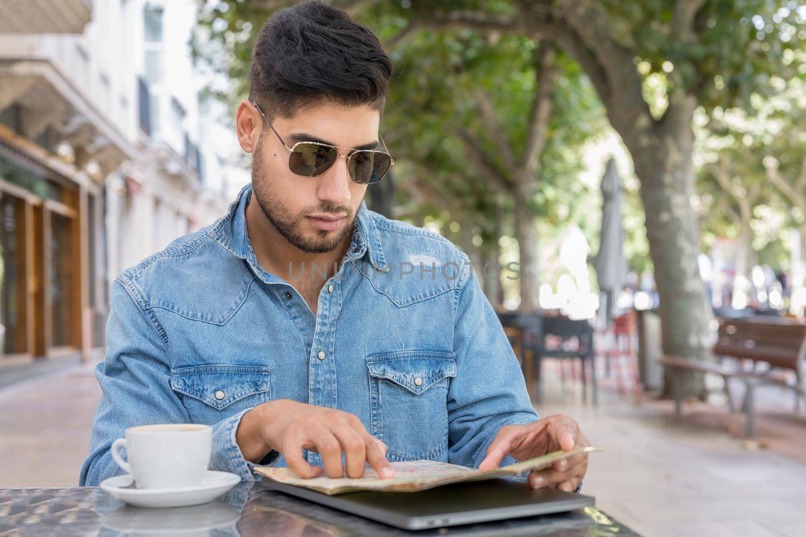 Young traveller man sitting in a cafe terrace and planning her trip with map and laptop by HERRAEZ