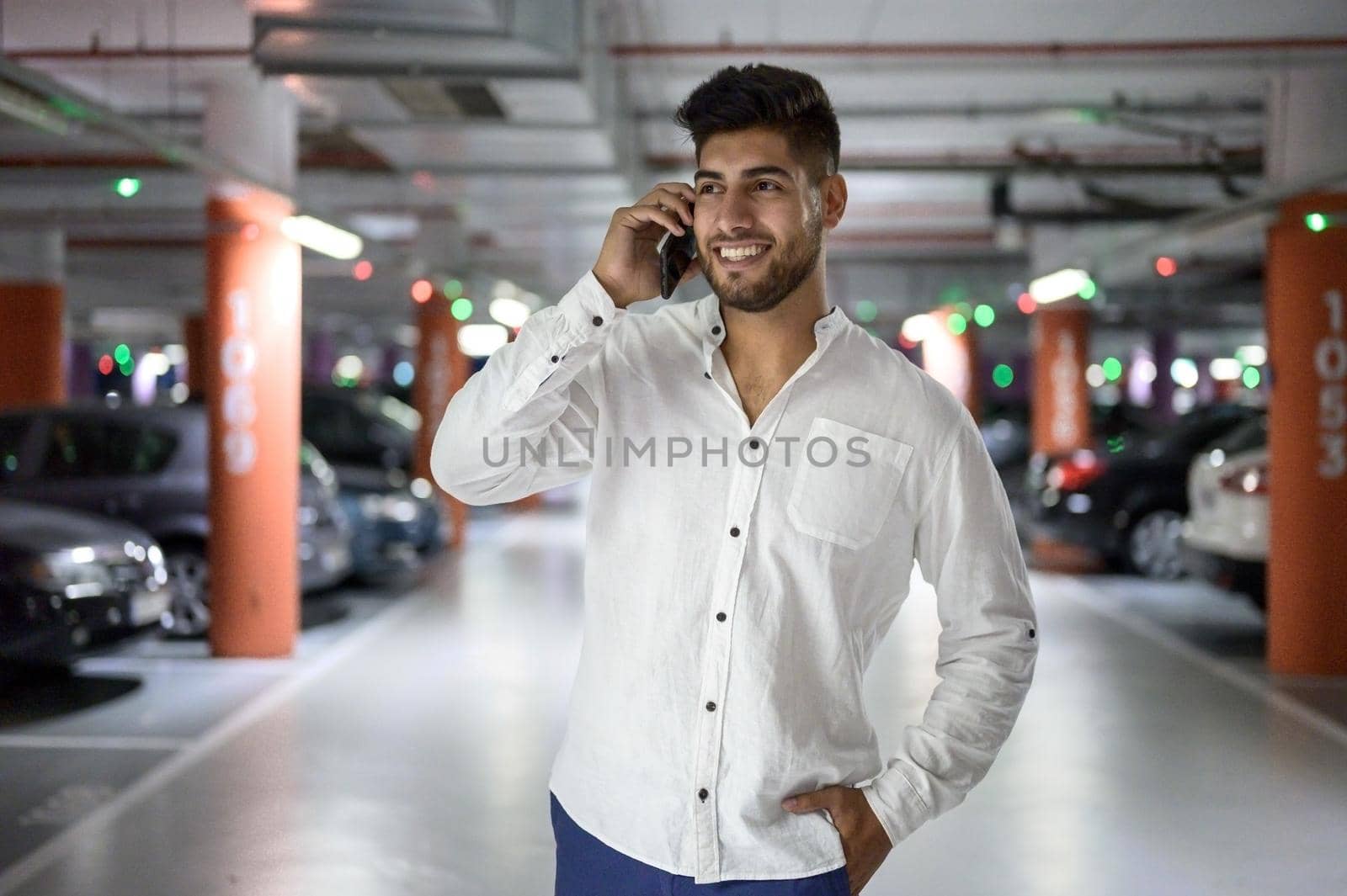 Confident, young handsome man walking along the parking lot while talking on the phone after having parked his car. High quality photo.