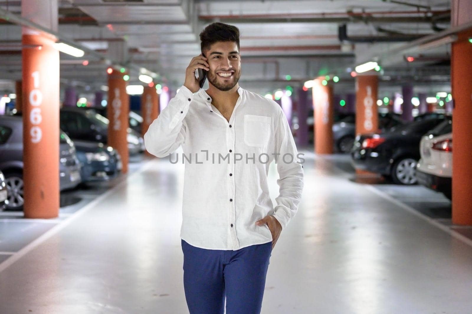 Confident, young handsome man walking along the parking lot while talking on the phone after having parked his car. by HERRAEZ