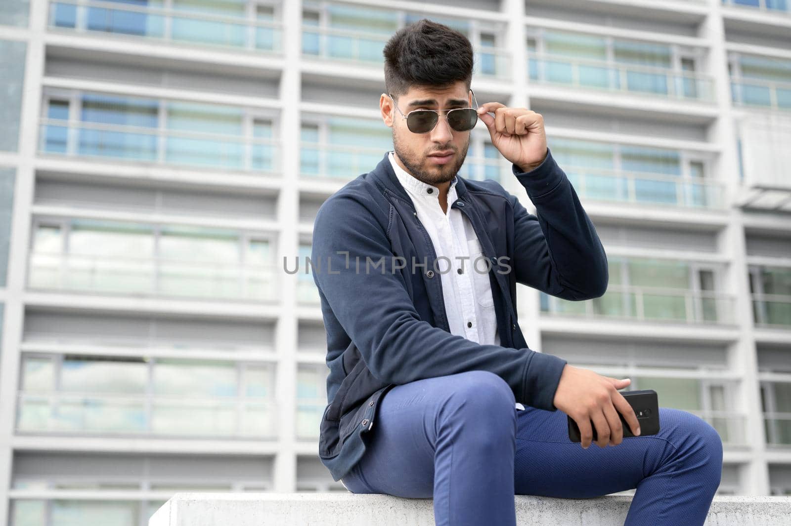 Portrait of young man in stylish sunglasses posing near modern building. by HERRAEZ