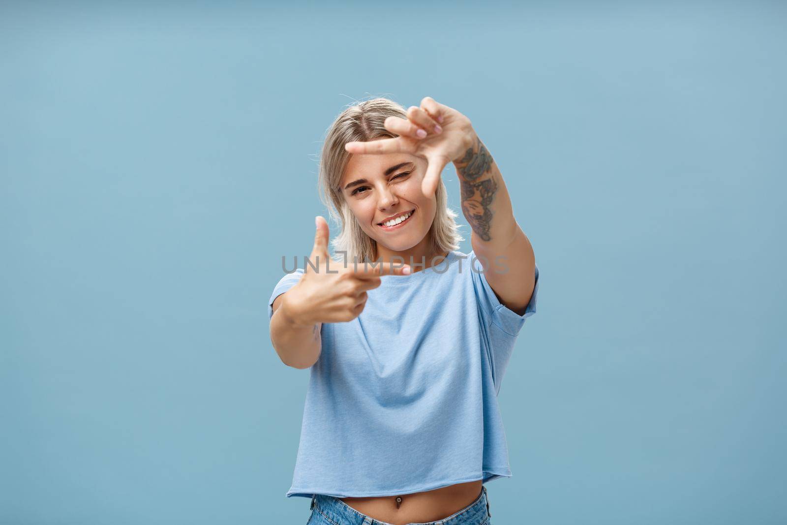 Be creative and happy. Portrait of charming enthusiastic female designer with blond hair and tattoo on arm winking and smiling joyfully making frame gesture as if measuring space over blue background.