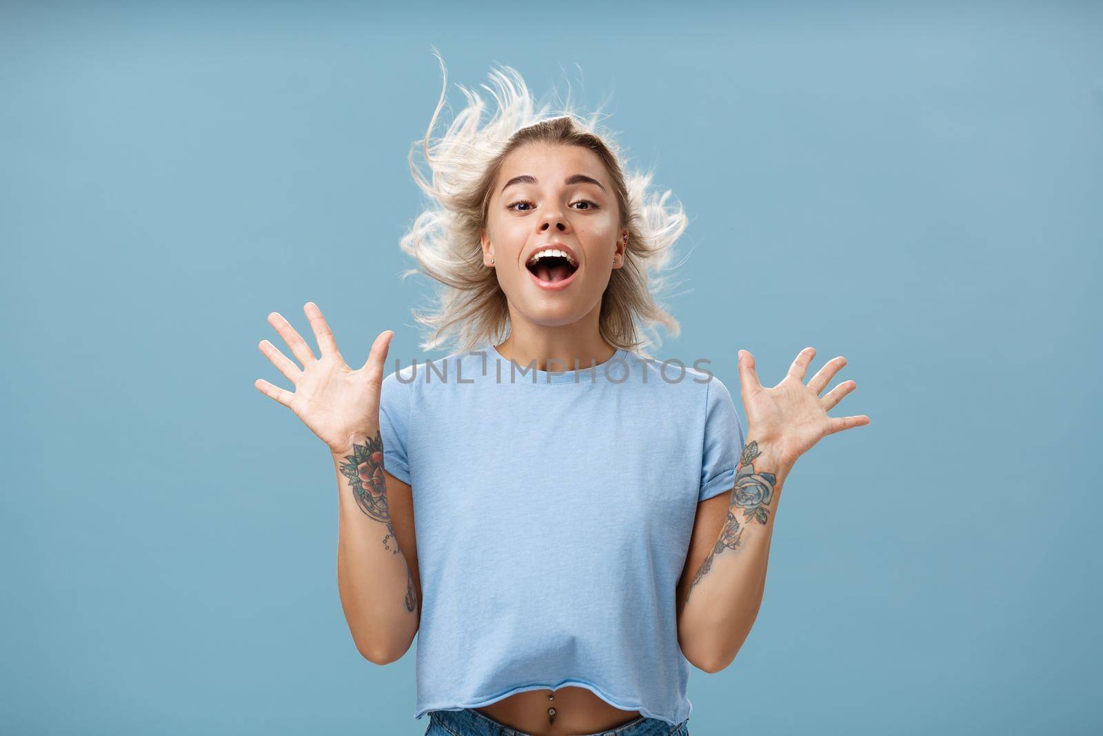 Creative happy and playful beautiful blond girl with tattoos on arms raising palms up opening mouth facing wind while hair strands flicking on air jumping having fun over blue background by Benzoix