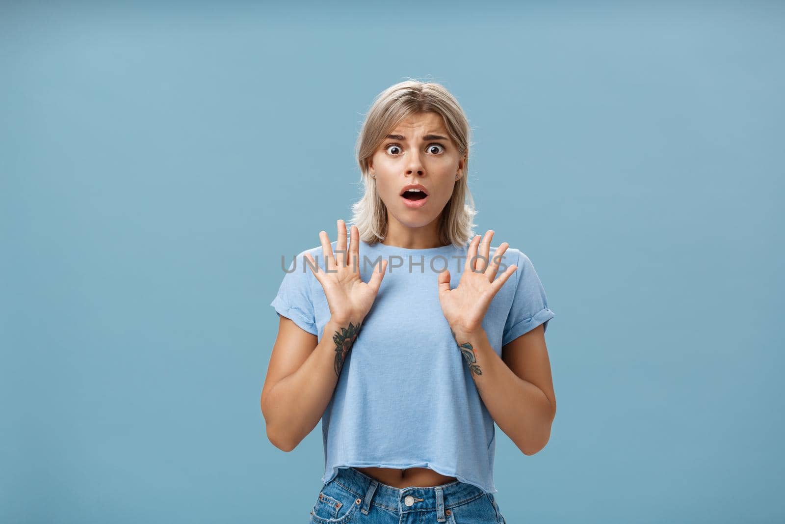 Shocked and worried concerned good-looking feminine woman with blond hair and tattooed arms raising palms gasping and opening mouth expressing empathy and feeling sorry for friend over blue wall. Emotions concept