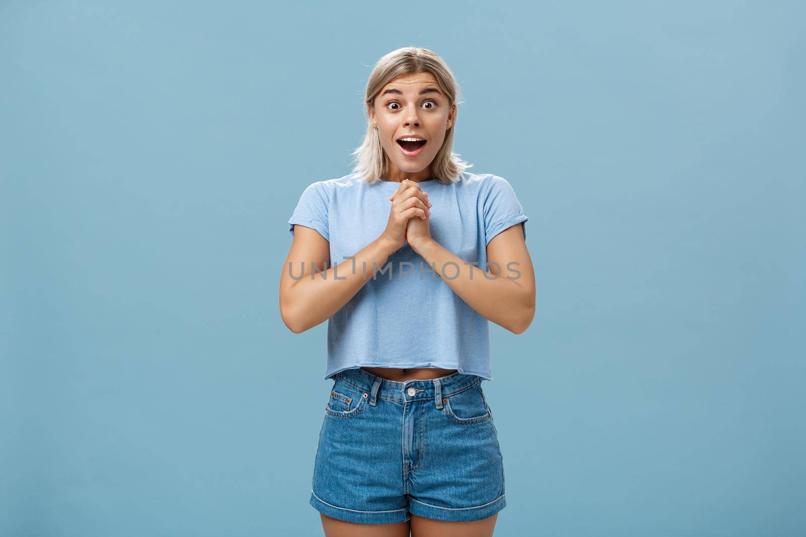 Lifestyle. Surprised girl receiving bouquet of flowers from secret fan holding hands together on breast gasping opening mouth from amazement and happiness popping eyes from astonishment over blue wall.