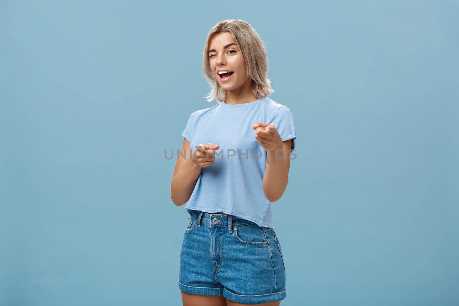 Hey I got offer for you. Portrait of attractive self-assured and playful stylish blond female with tanned skin in denim shorts and summer t-shirt winking and pointing at camera over blue background by Benzoix