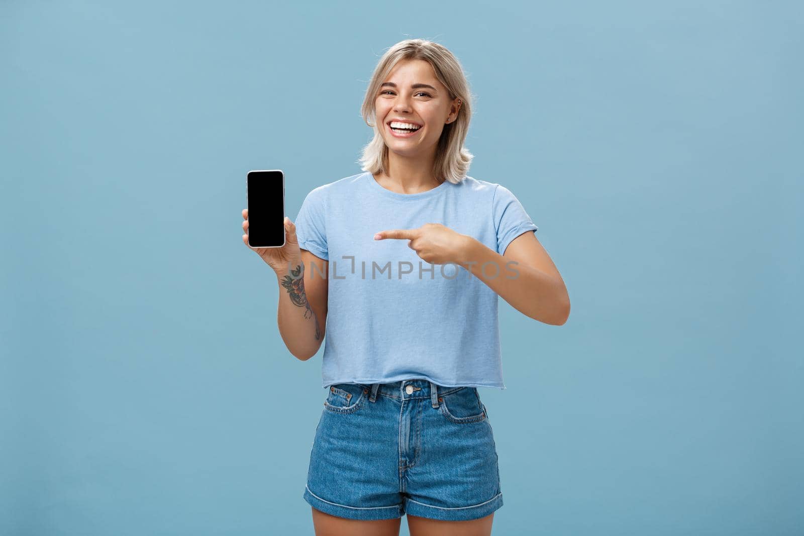 Look at this hilarious photo. Entertained attractive happy woman with fair hair in casual t-shirt and denim shorts showing smartphone at camera pointing at device screen smiling broadly over blue wall by Benzoix