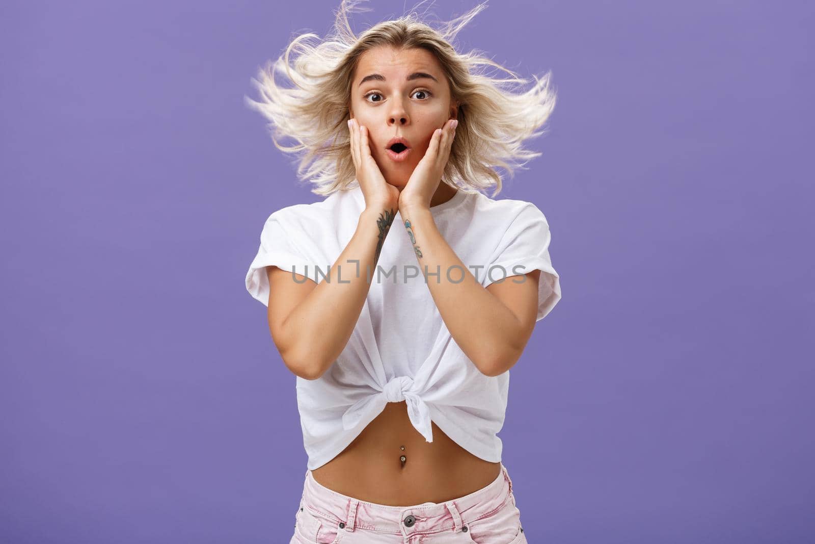 Romantic surprised and thrilled feminine blonde female with tattoos standing on windy street folding lips and holding hands on face impressed and intrigued while hair floats in air over purple wall by Benzoix
