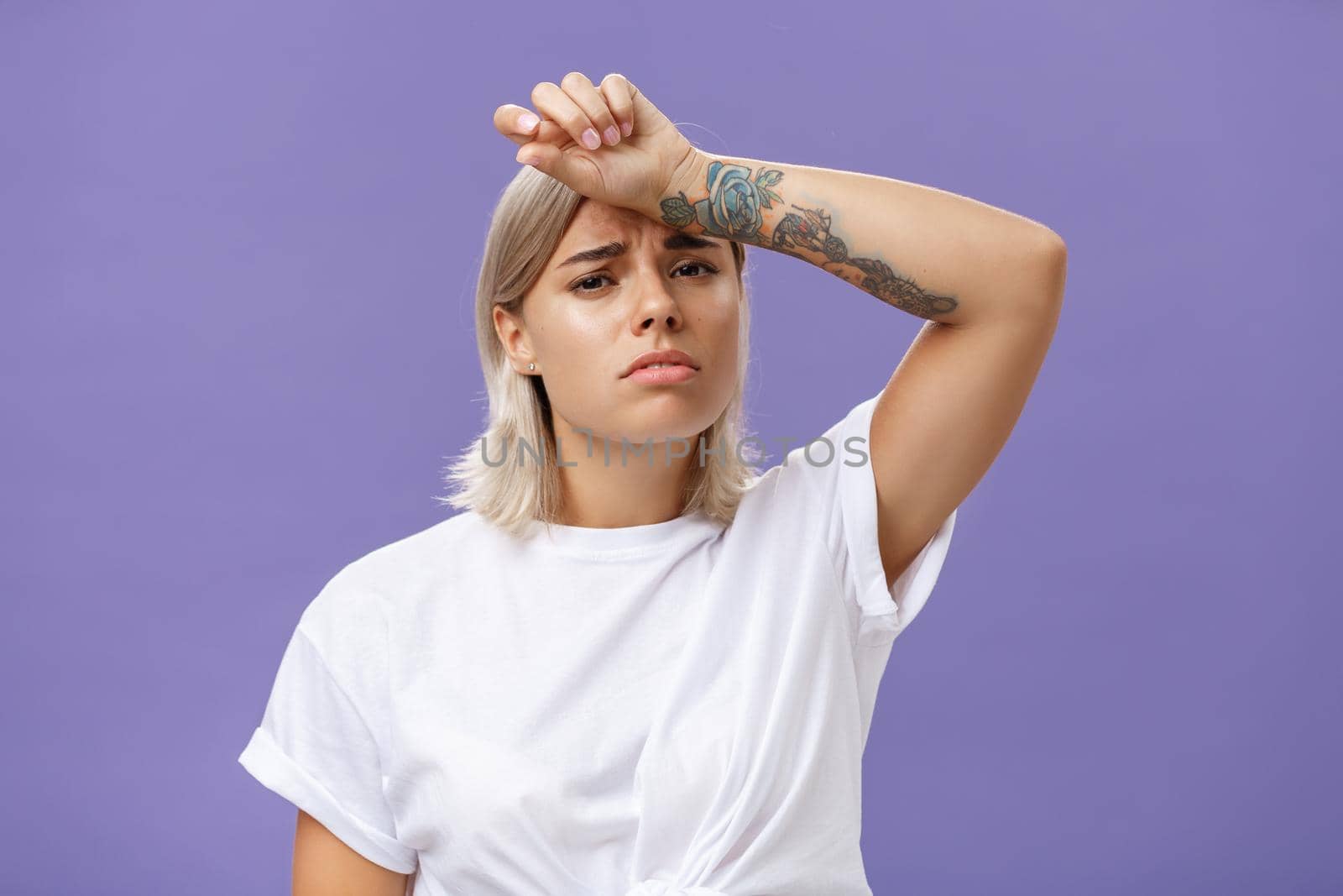 Indoor shot of tired and displeased exhausted cute european woman with tanned skin and tattoos on arms holding hand on forehead frowning suffering from tiresome work or sun heating over purple wall by Benzoix