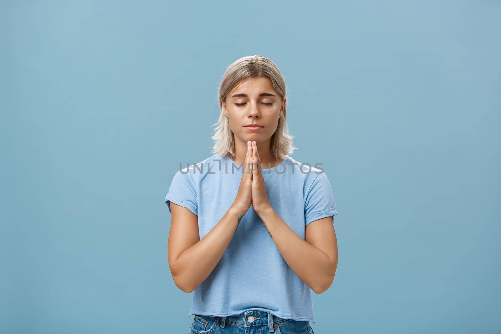 Lifestyle. Good-looking kind and faithful european girl with blond hair smiling cute and tender holding hands in pray while making wish hopefully believing god hearing her prayers with closed eyes over blue wall.