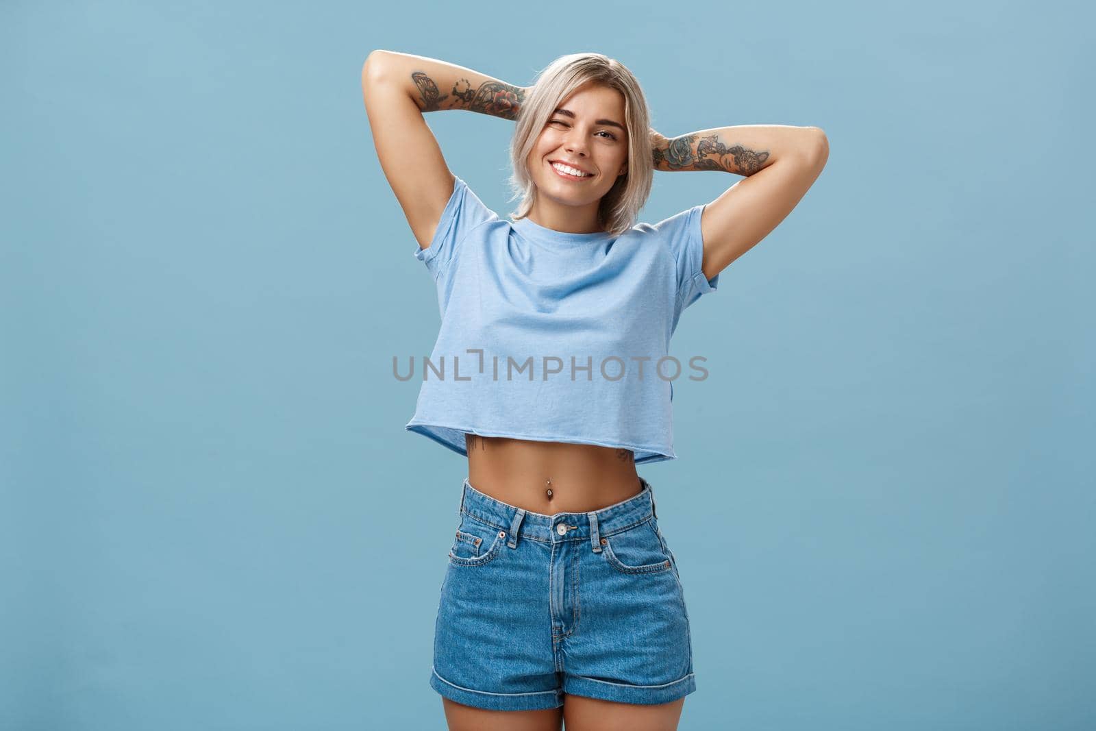 Thanks god it weekends. Relaxed lazy and attractive young stylish woman with fari hair tattoos, pierced belly stretching with delight holding hands behind head in carefree pose, smiling over blue wall.