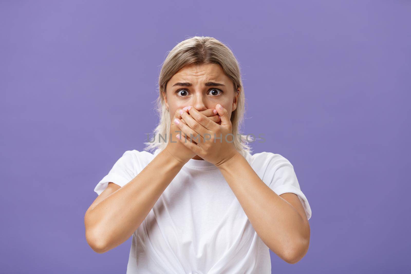 Waist-up shot of shocked nervous and scared young woman witnessing terrible crime covering mouth with both hands not to scream frowning staring frightened at camera over purple background by Benzoix