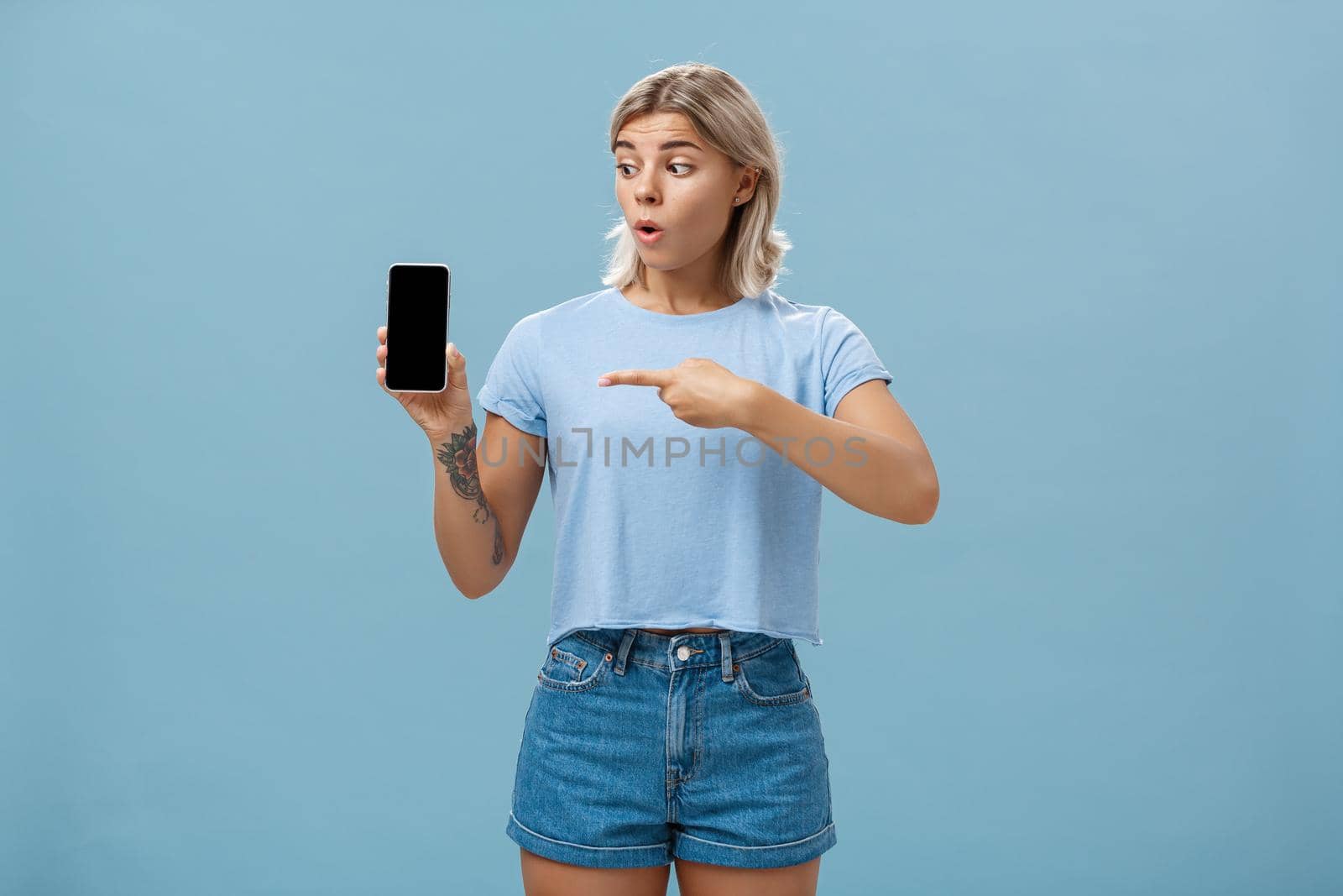 You should download app. Portrait of amazed shocked attractive blonde girl in trendy summer t-shirt holding smartphone gasping from surprise pointing at gadget screen posing over blue background by Benzoix