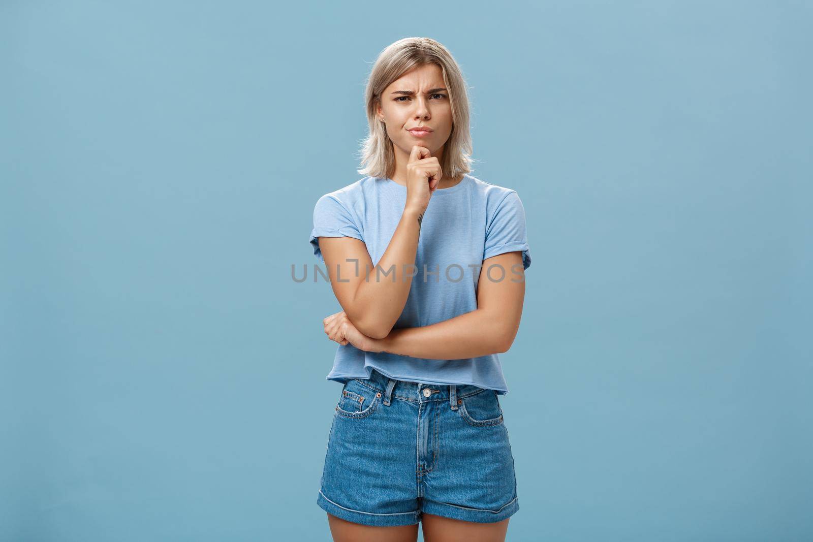 I doubt it good idea. Suspicious intense creative young female coworker in outdoor outfit frowning from doubtful thoughts holding hand on chin while thinking expressing disbelief over blue wall by Benzoix