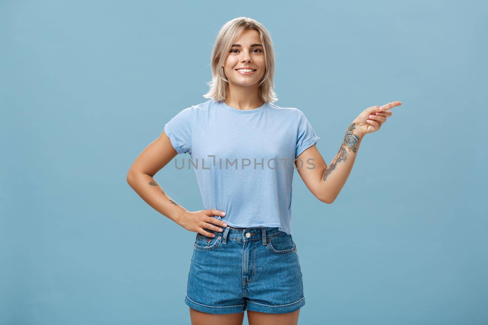 Check it out. Portrait of beautiful confident young caucasian girl with blond hair pointing right and holding hand on waist in self-assured relaxed pose smiling broadly over blue background by Benzoix
