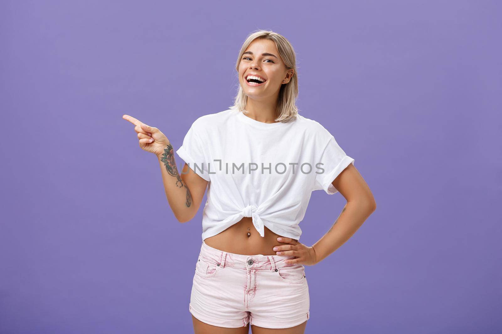 Check out cool place. Satisfied carefree stylish and attractive adult woman with perfect body tanned skin, tattoos laughing and smiling joyfully holding hand on hip and pointing left over purple wall.