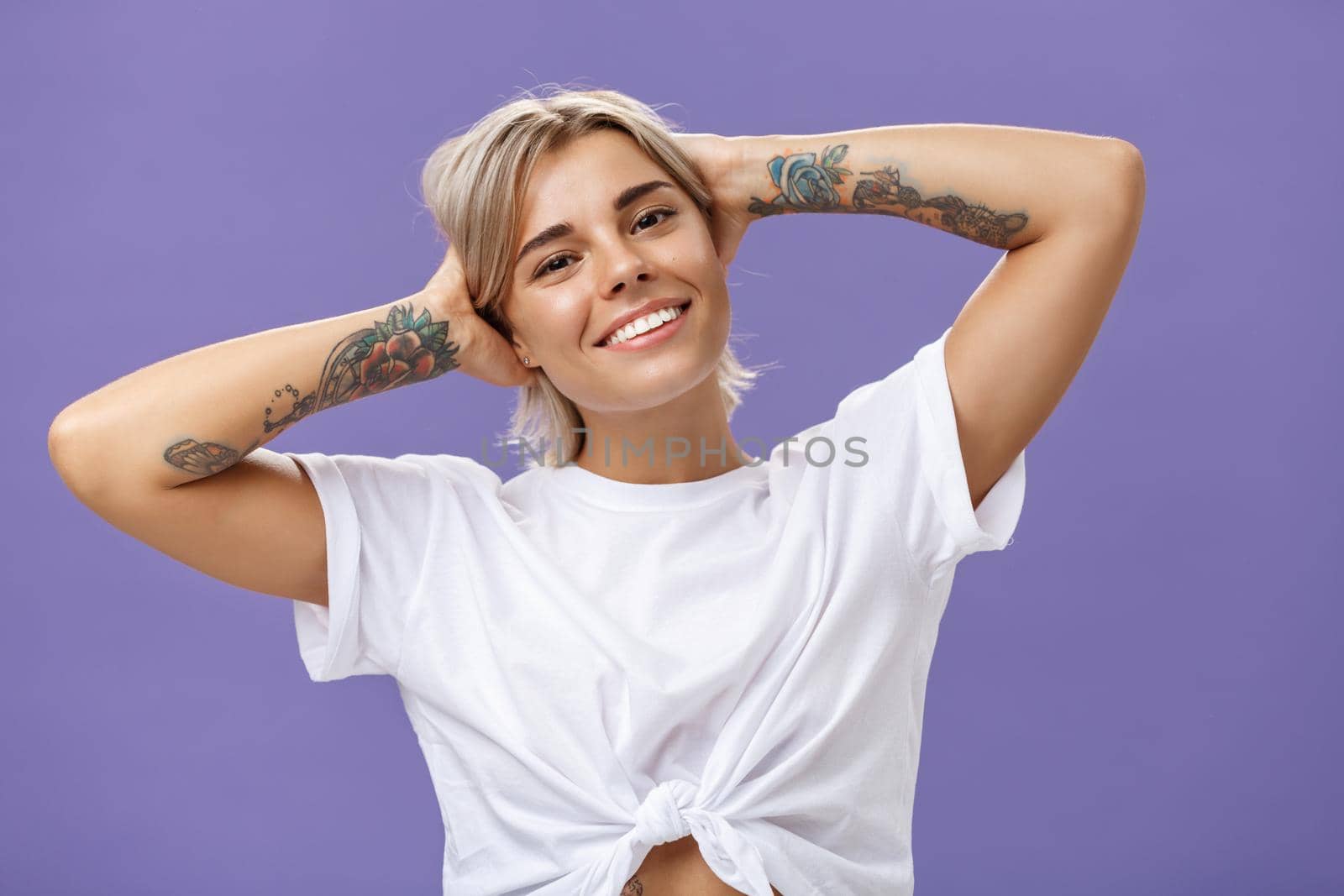 Lifestyle. Close-up shot of good-looking stylish and relaxed blond woman with tattoos on arms holding hands behind head smiling with carefree pleased look enjoying weekends over purple background.