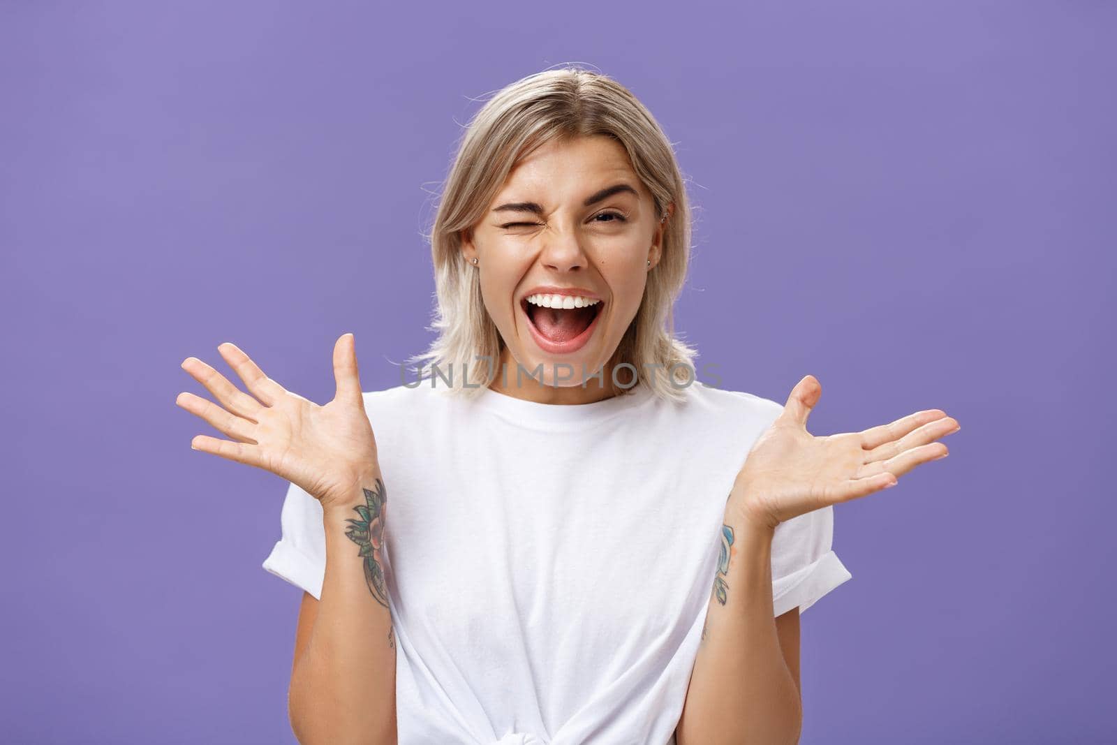 Waist-up shot of playful energized sociable attractive woman with blond hair, tanned skin and tattoos raising hands joyfully and spread aside winking from amusement smiling broadly over purple wall.