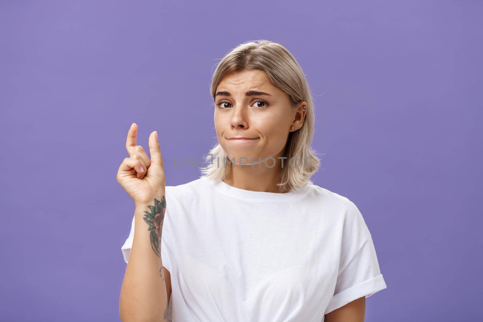 Girl having tiny problem. Concerned attractive blonde girl with tattoo on arm pursing lips in troubled look shaping small or little object, dissatisfied with regret in eyes over purple wall. Copy space