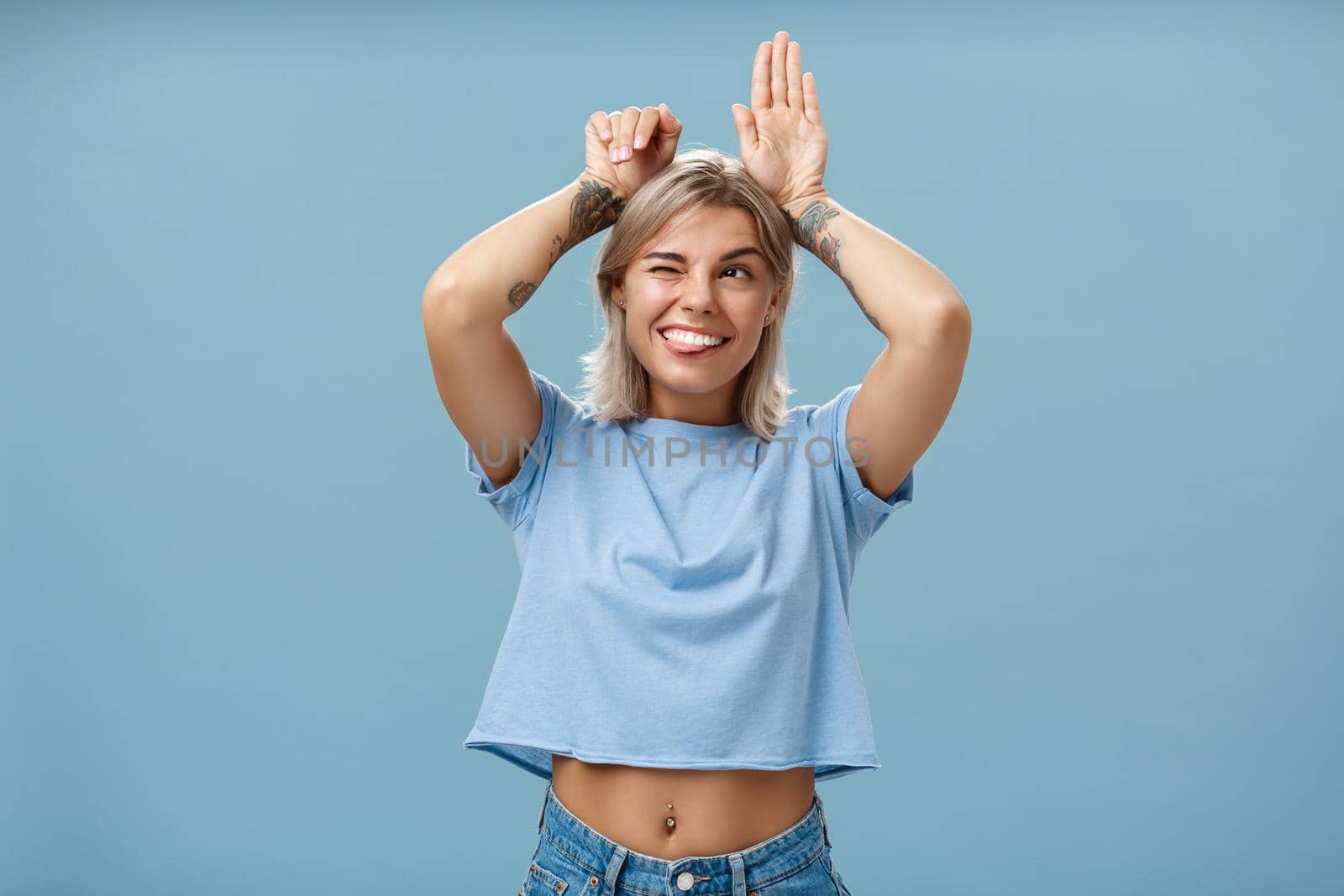 Studio shot of entertained carefree and emotive happy charming woman with tattoos on arms acting like bunny with palms on head winking joyfully smiling and sticking out tongue over blue wall by Benzoix