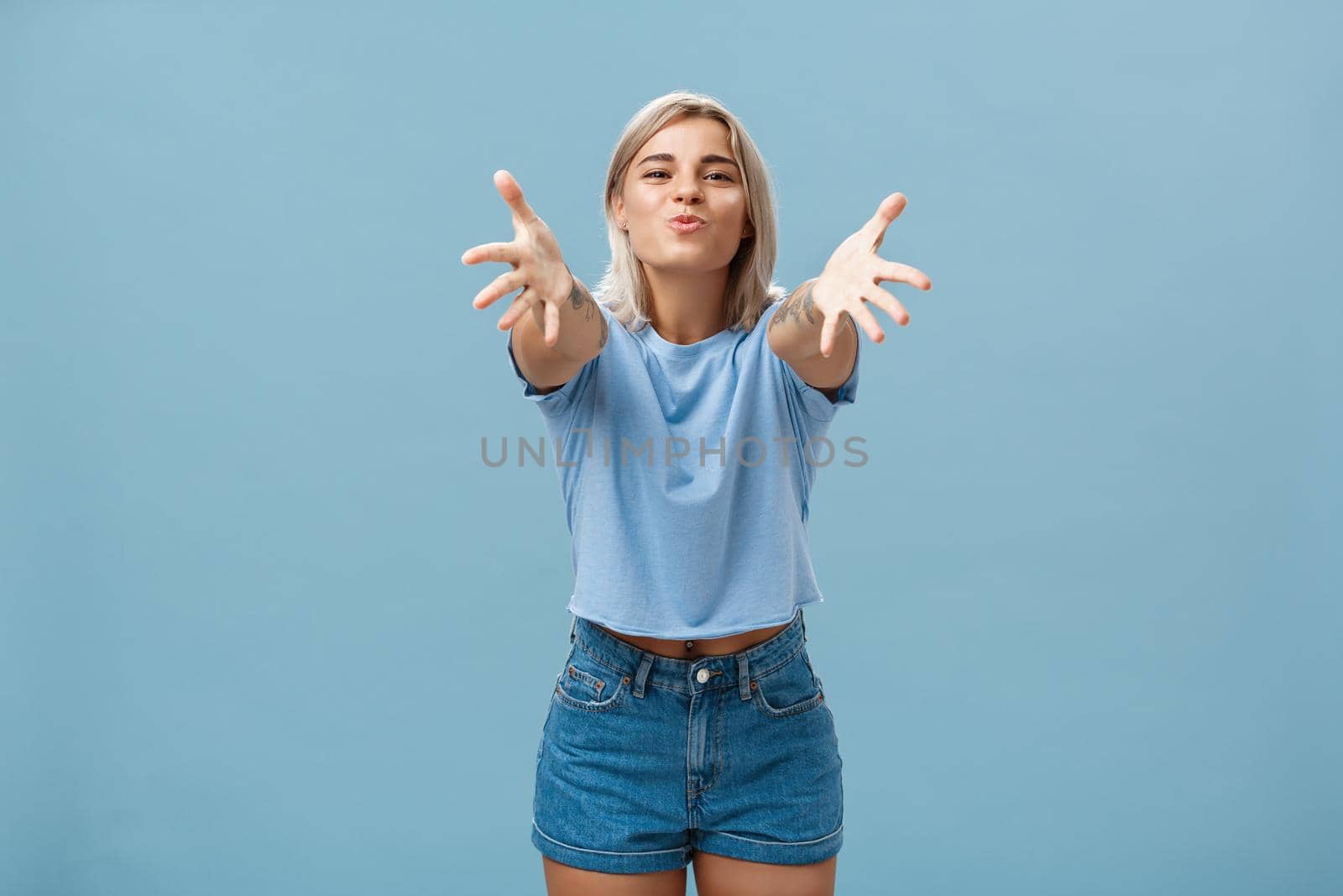 Come to mama. Portrait of friendly joyful and enthusiastic caucasian female student in trendy outfit pulling hands towads camera and folding lips to hug and give passionate kiss smiling over blue wall by Benzoix
