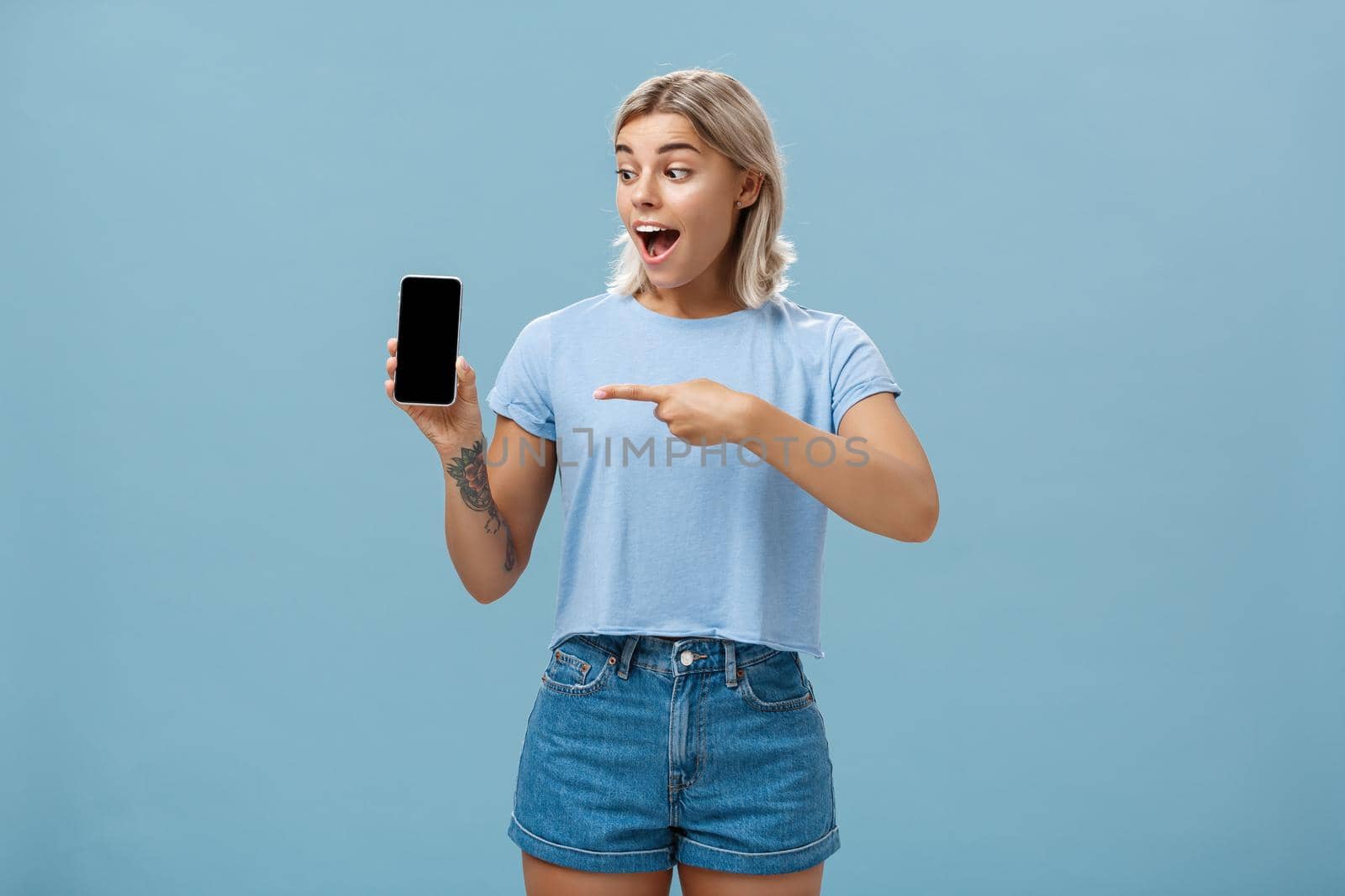 Look at this awesome smartphone. Impressed enthusiastic good-looking female blonde with cool tattoo on arm dropping jaw from thrill and excitement pointing at device screen over blue background by Benzoix