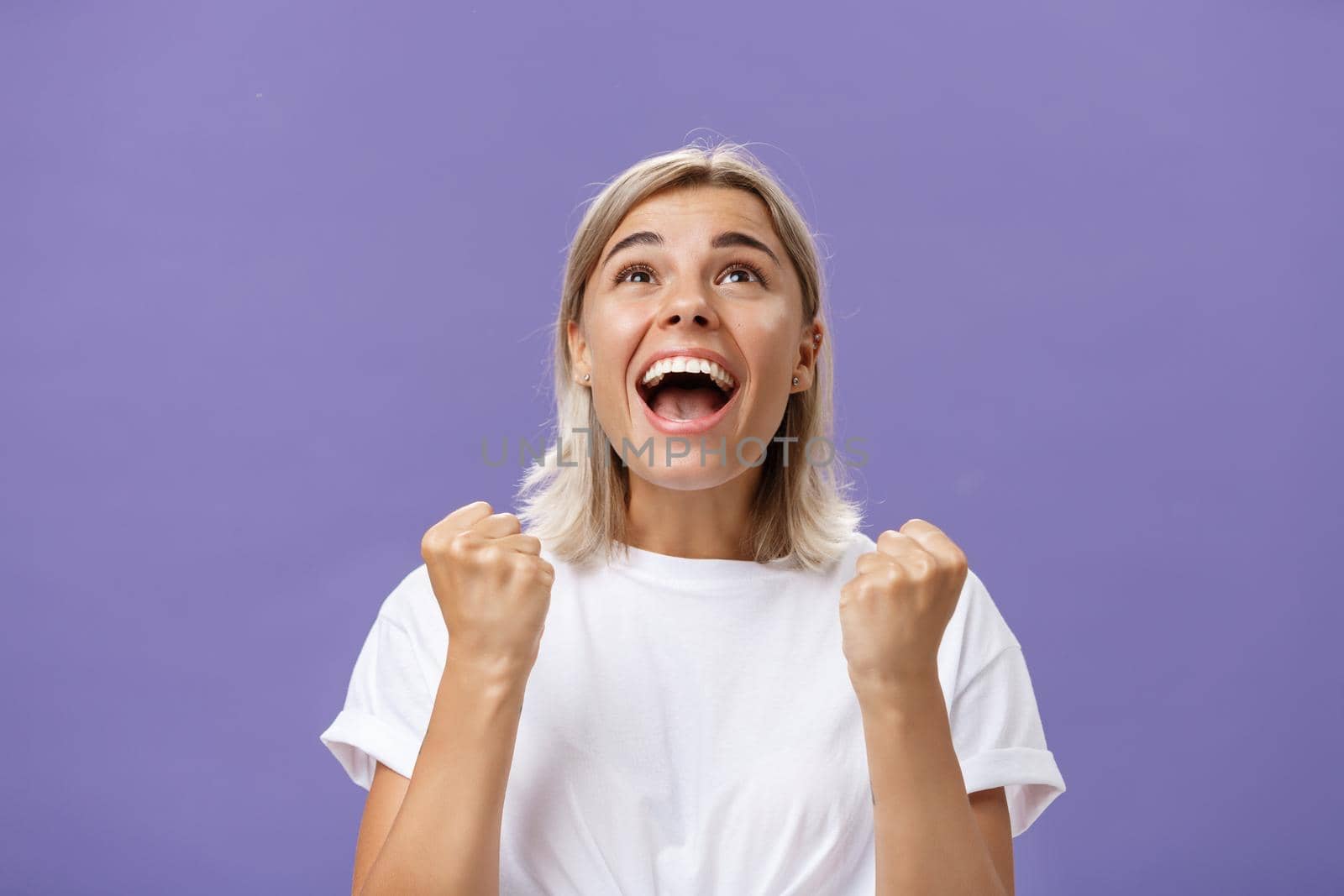 Waist-up shot of grateful delighted lucky girl with attractive tan in white t-shirt clenching fist from positive emotions gazing delighted and happy up thankful giving thanks god for fulfilling dream by Benzoix