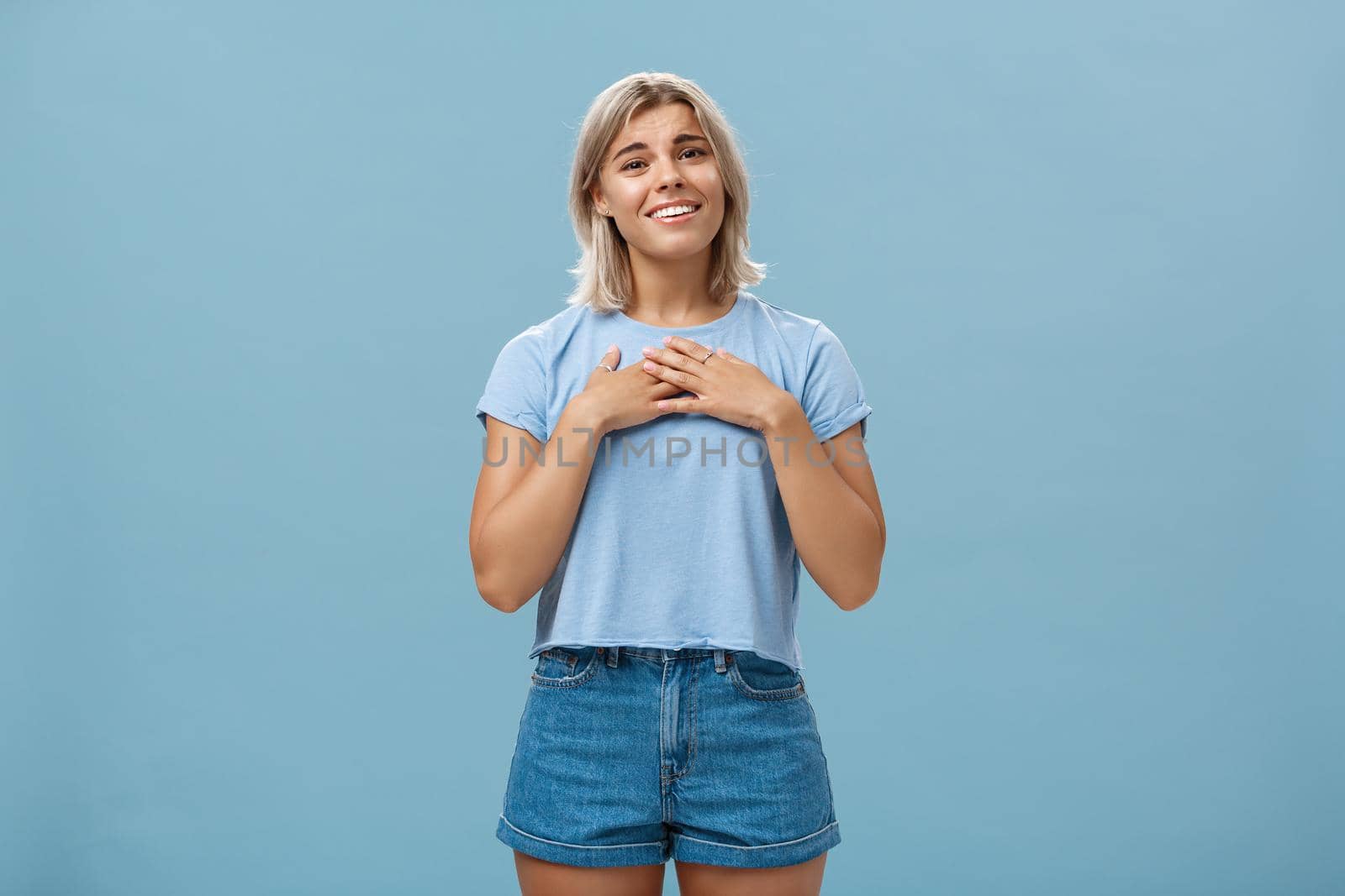 So sweet thanks. Touched beautiful and emotive blond girl in t-shirt and shorts with tanned skin holding palms on breast, sighing and smiling joyfully being thankful and grateful over blue wall. Emotions concept