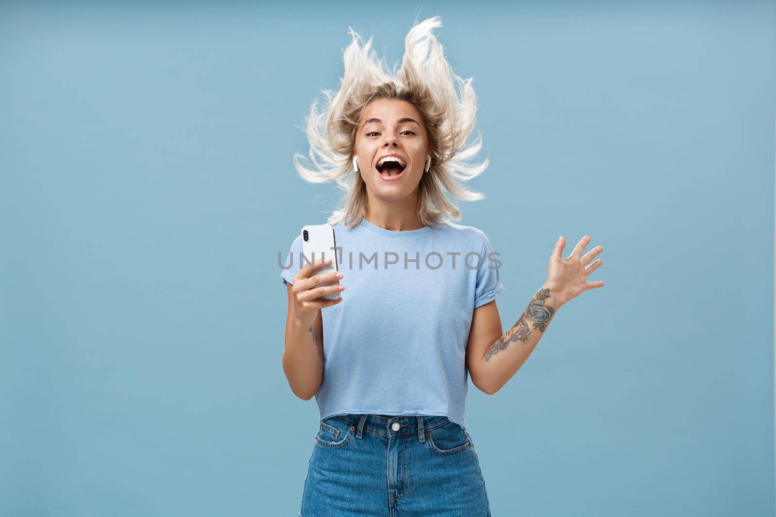 Expressing happiness with help of great tunes. Joyful amused and happy good-looking young female student jumping having fun listenign music in wireless earbuds, holding smartphone over blue wall.