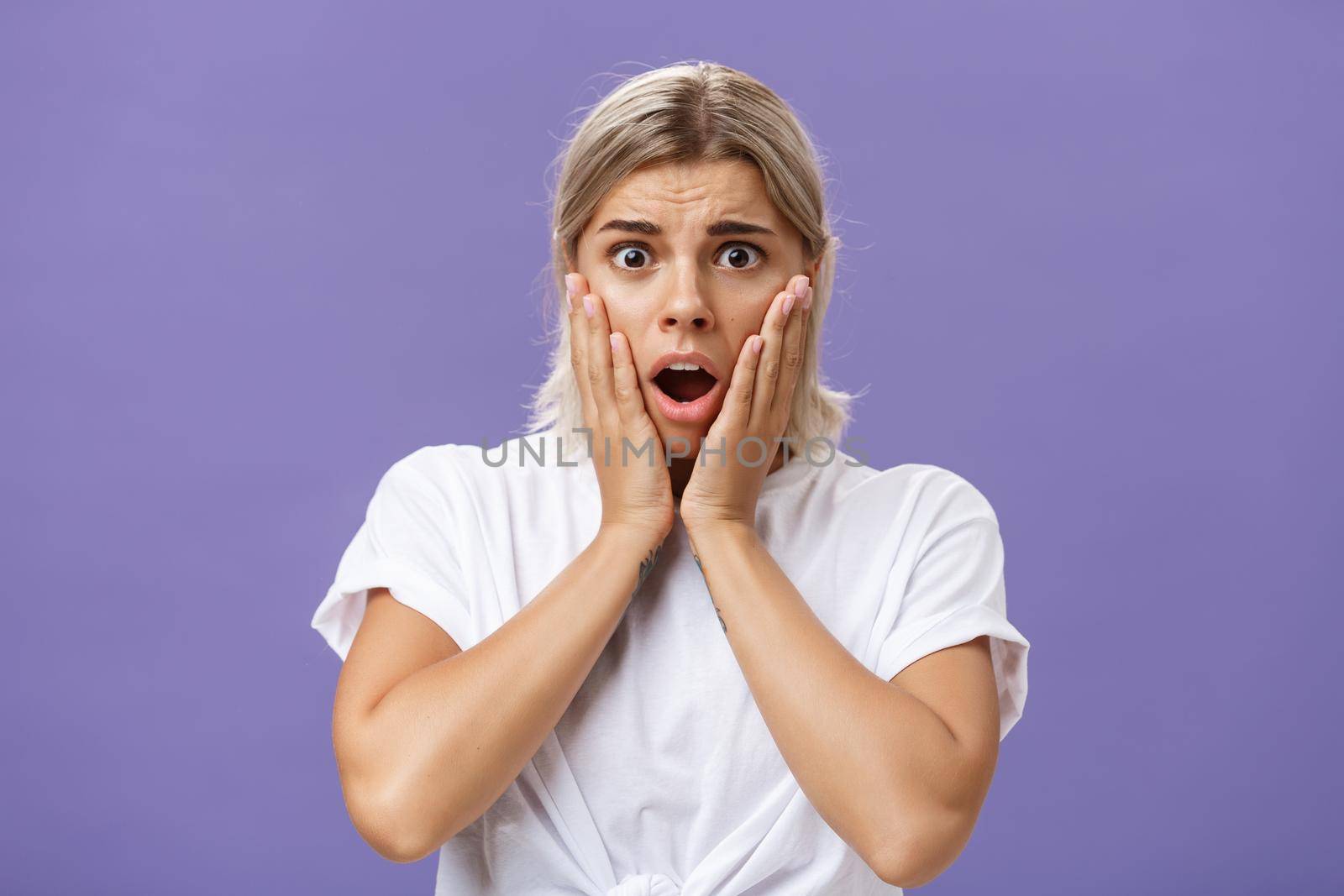 Shocked and worried silly european woman expressing empathy feeling sorry for poor animal in danger gasping opening mouth holding hands on face frowning feeling troubled over purple wall by Benzoix