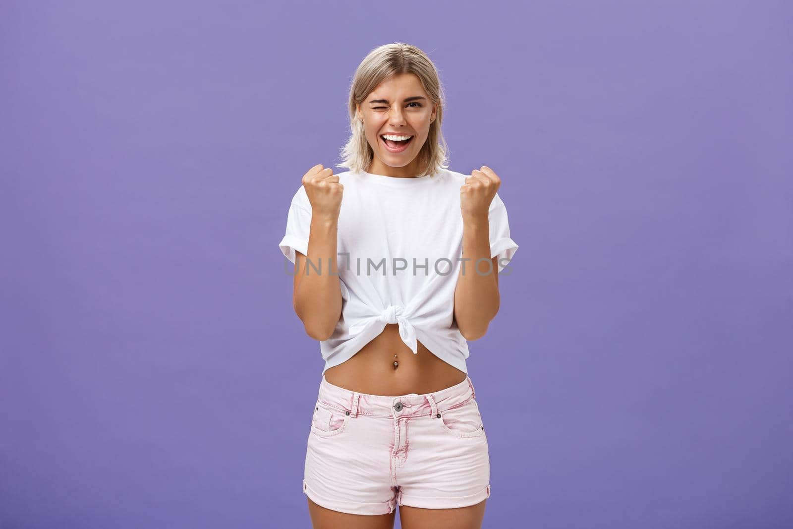 Rejoicing satisfied and pleased happy young ambitious girl with blonde hair in trendy white cropped top and shorts saying yes winking and smiling clenching fists in success or victory sign. Emotions and body language concept