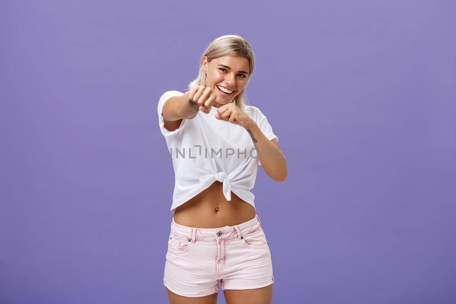 Ready fight back. Energized happy and awesome blonde female with tanned skin and fit body pulling fist in attack gesture towards camera smiling joyful acting like fighting or boxing over purple wall by Benzoix