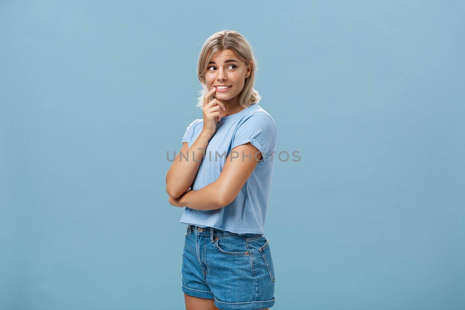Indoor shot of feminine romantic and timid good-looking european female with fair hair holding finger on lip smiling curious and intrigued while turning right and observing interesting thing. Copy space